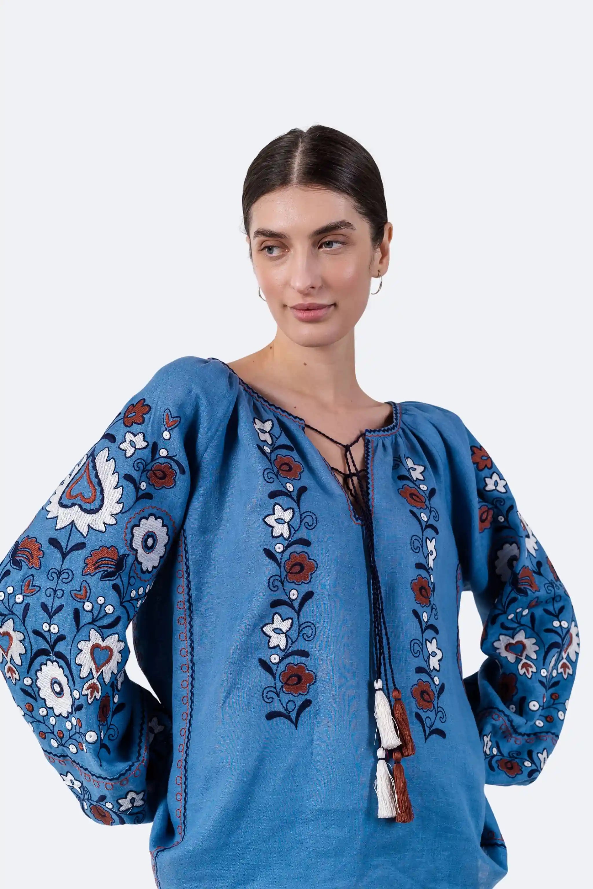 A woman modeling a blue embroidered blouse with floral patterns and a lace-up neckline, photographed against a plain light gray studio background.