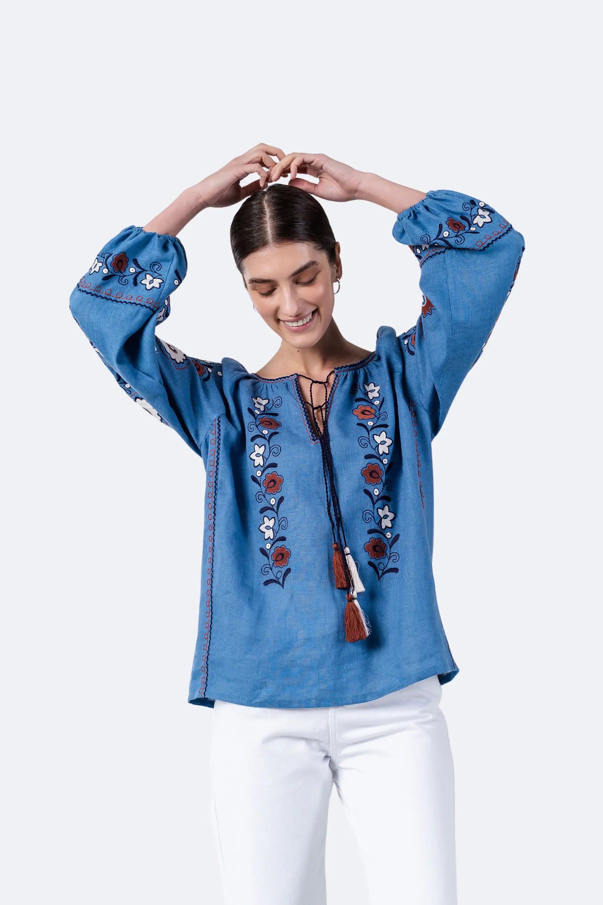 A woman wearing a blue long-sleeve embroidered blouse with white pants, posing against a light gray background.