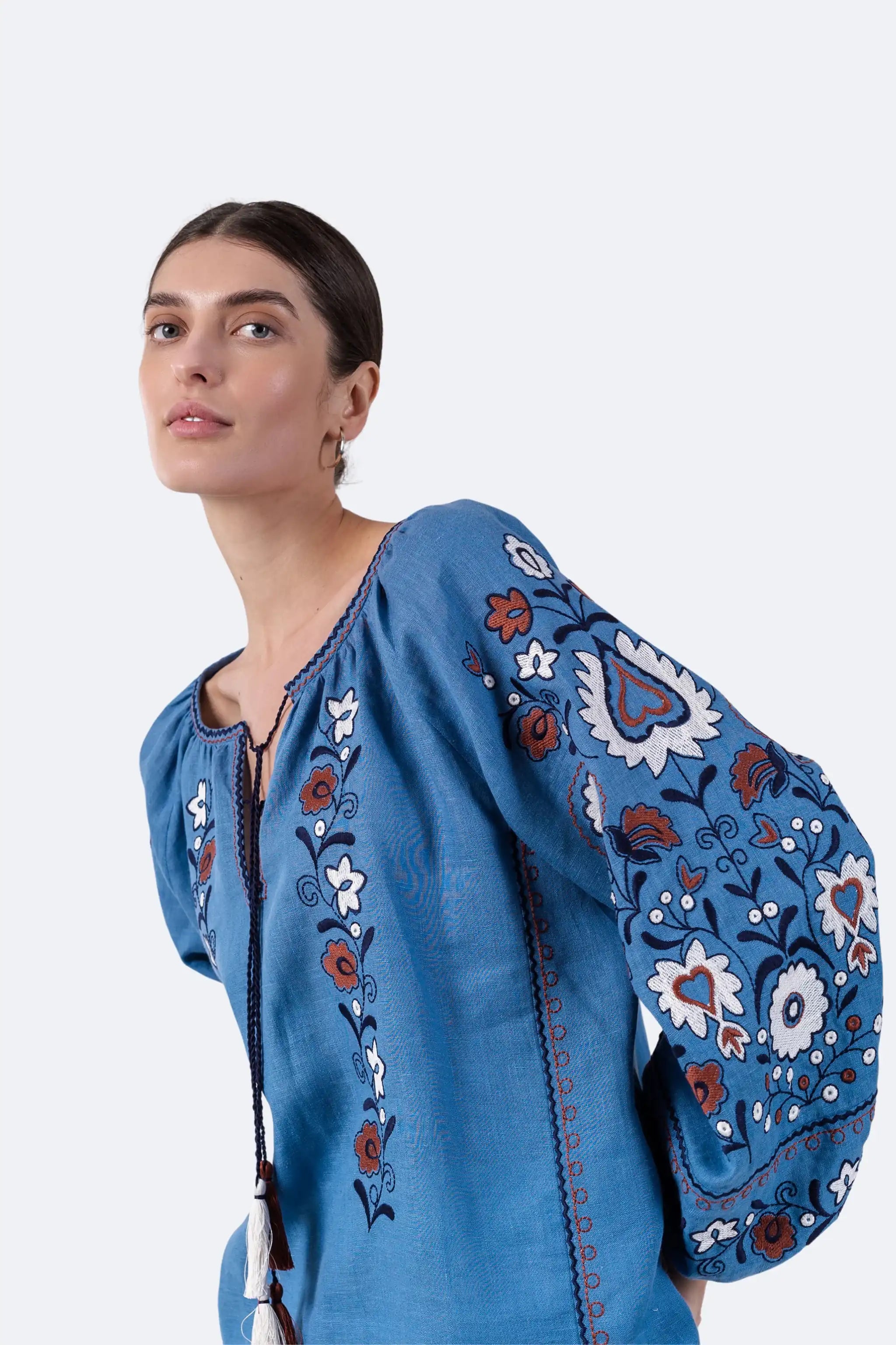 A woman wearing a blue embroidered blouse with floral and paisley patterns in white, red, and brown, photographed against a plain light gray studio background.