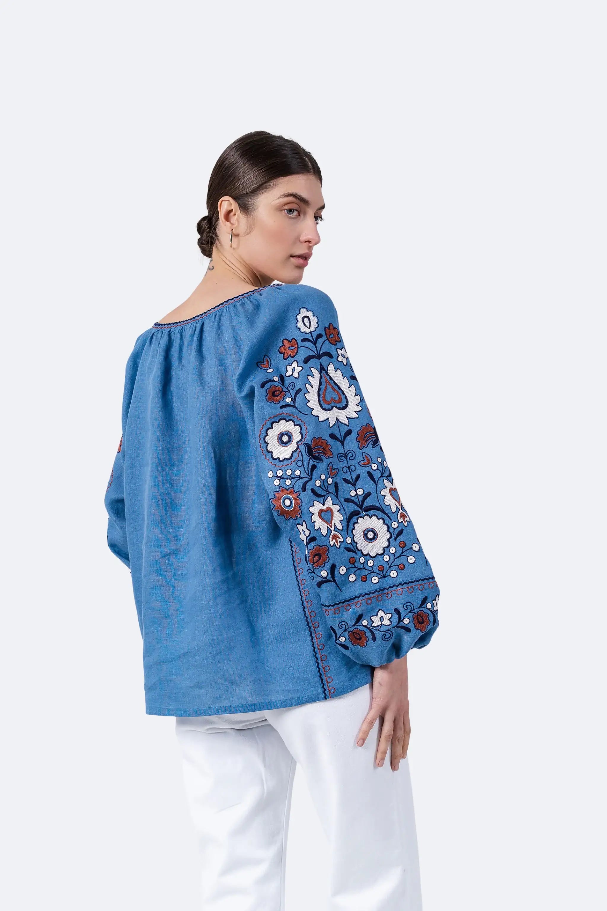 A woman wearing a blue embroidered blouse with white pants, photographed against a clean light gray studio background. The blouse has a wide neckline and long sleeves with detailed floral and paisley embroidery in blue, white, red, and brown.