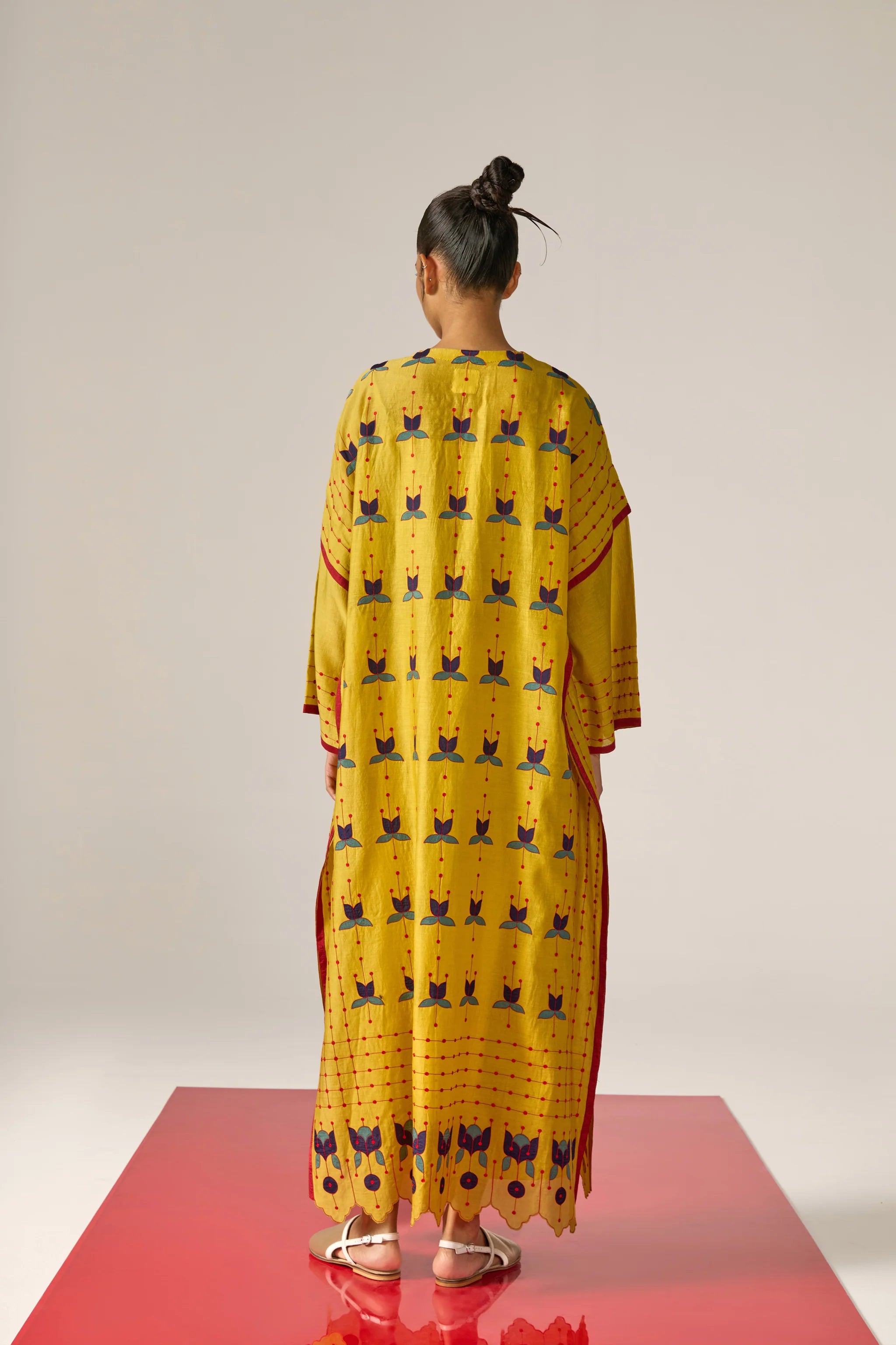 A model in a long mustard yellow kaftan with blue floral appliqué, red threadwork grid patterns, and a scalloped cutwork hem.