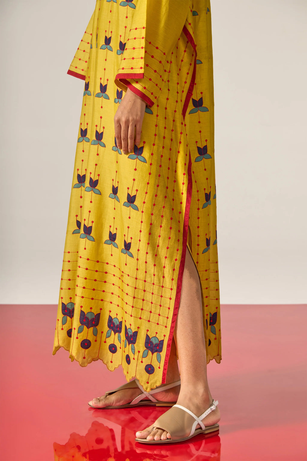 A model in a long mustard yellow kaftan with blue floral appliqué, red threadwork grid patterns, and a scalloped cutwork hem.