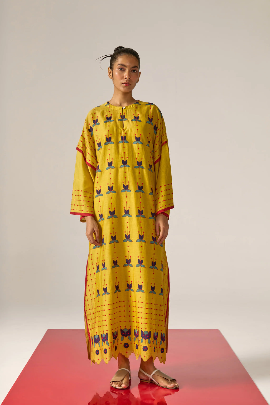 A model in a long mustard yellow kaftan with blue floral appliqué, red threadwork grid patterns, and a scalloped cutwork hem.