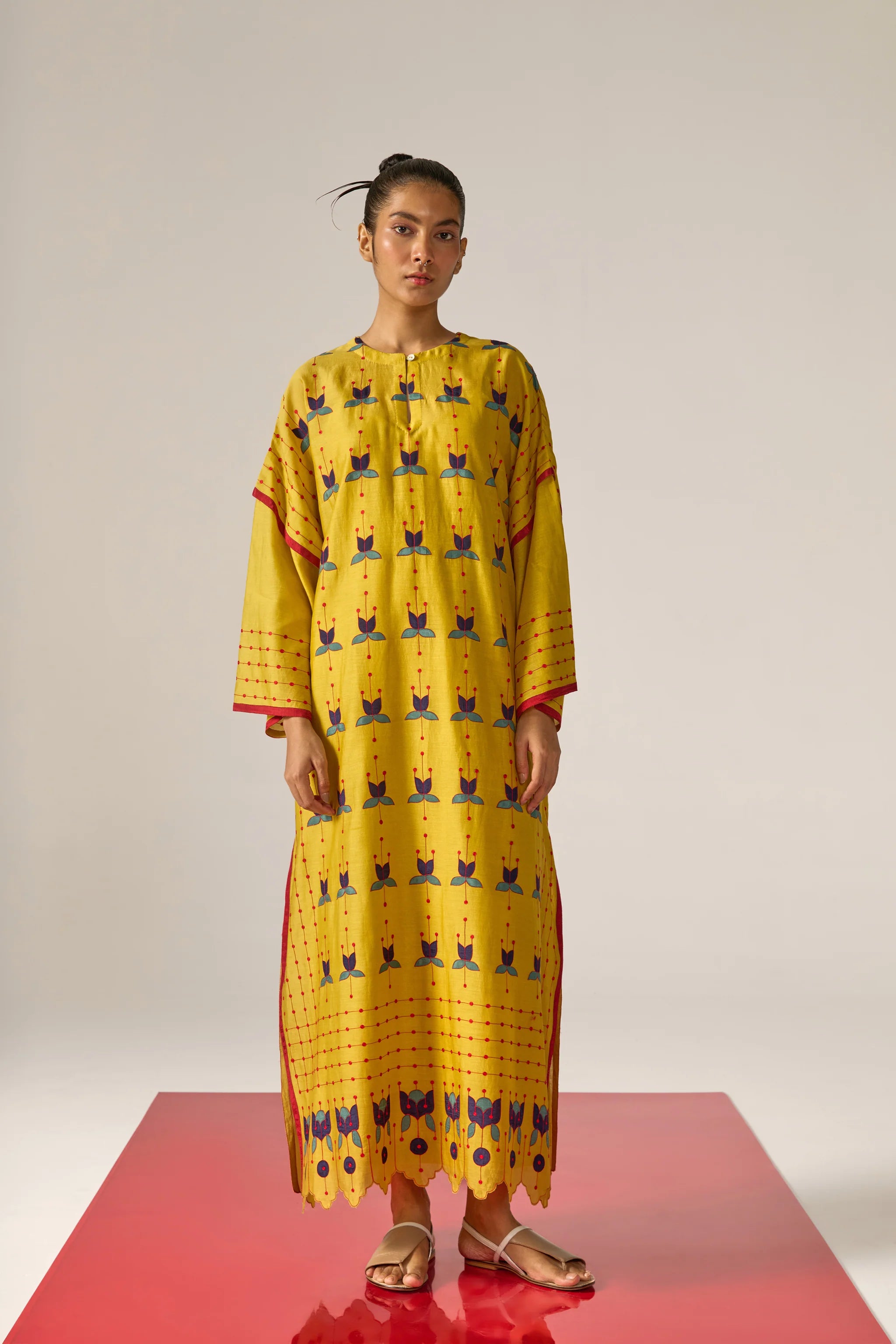 A model in a long mustard yellow kaftan with blue floral appliqué, red threadwork grid patterns, and a scalloped cutwork hem.