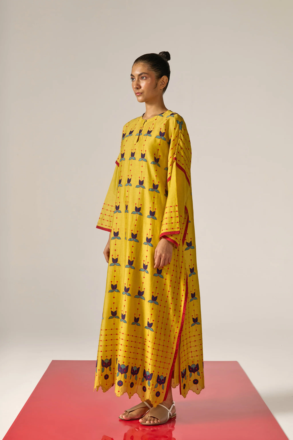 A model in a long mustard yellow kaftan with blue floral appliqué, red threadwork grid patterns, and a scalloped cutwork hem.