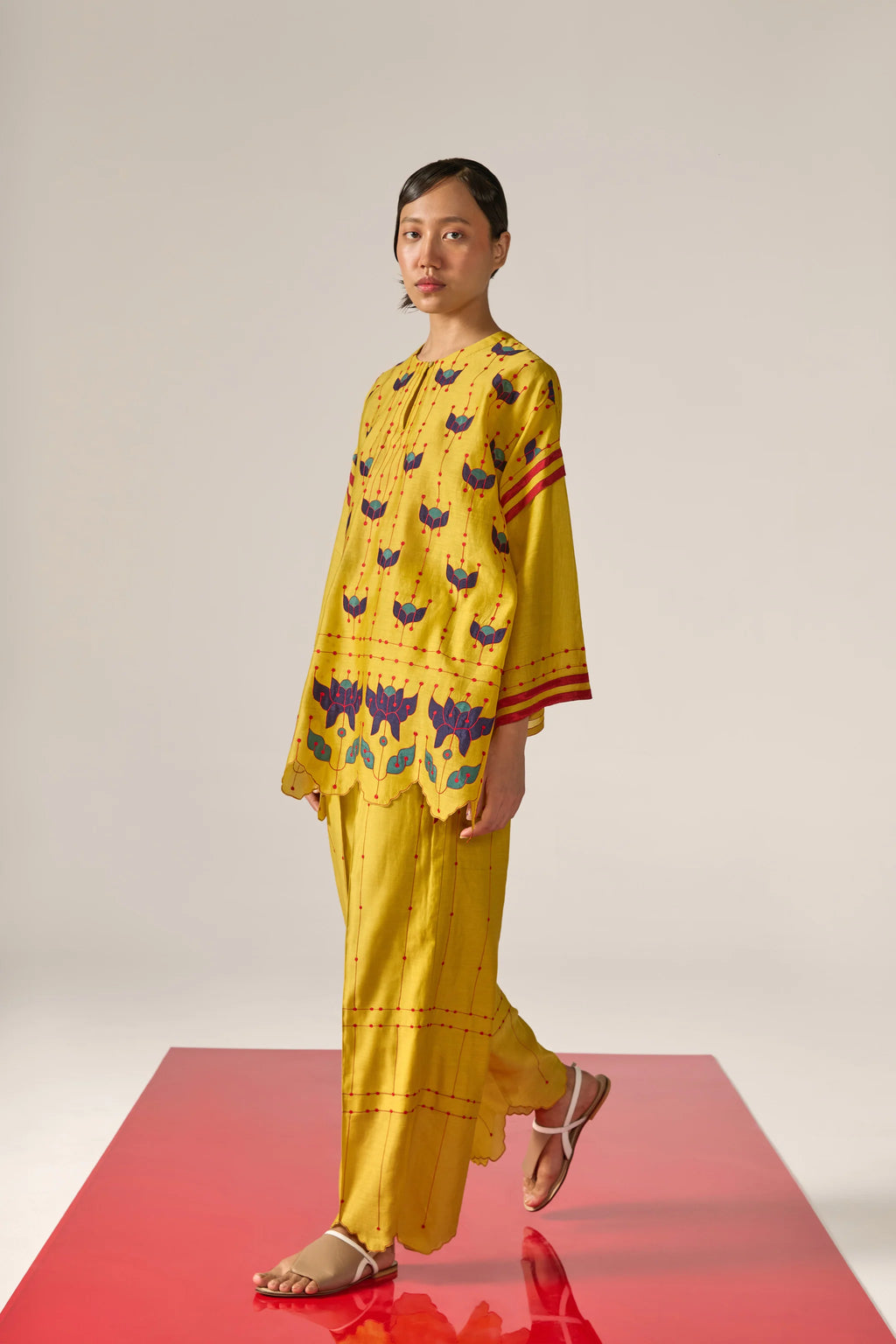 A  woman model wearing yellow Cotton Silk Chanderi kaftan shirt with teal geometric appliqué and cutwork detailing on the sleeves and back, paired with matching trousers.