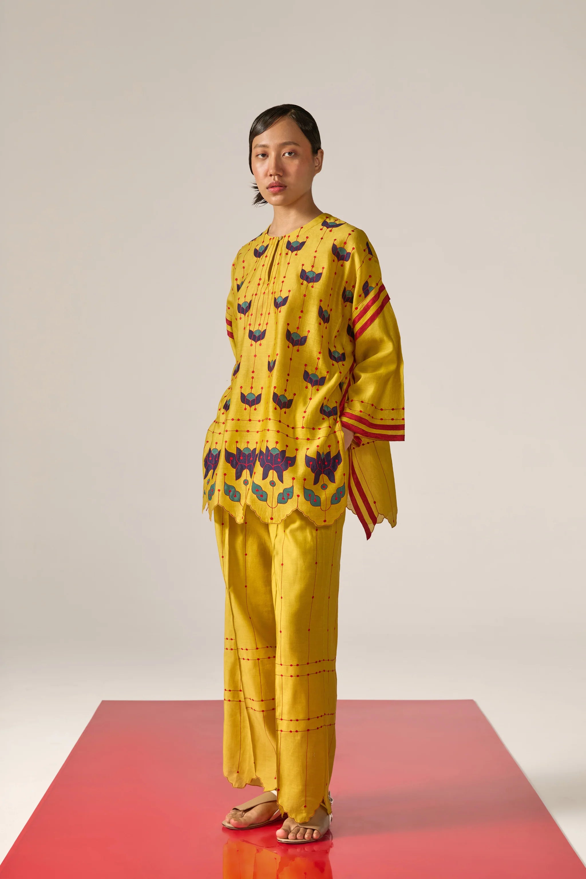 A  woman model wearing yellow Cotton Silk Chanderi kaftan shirt with teal geometric appliqué and cutwork detailing on the sleeves and back, paired with matching trousers.