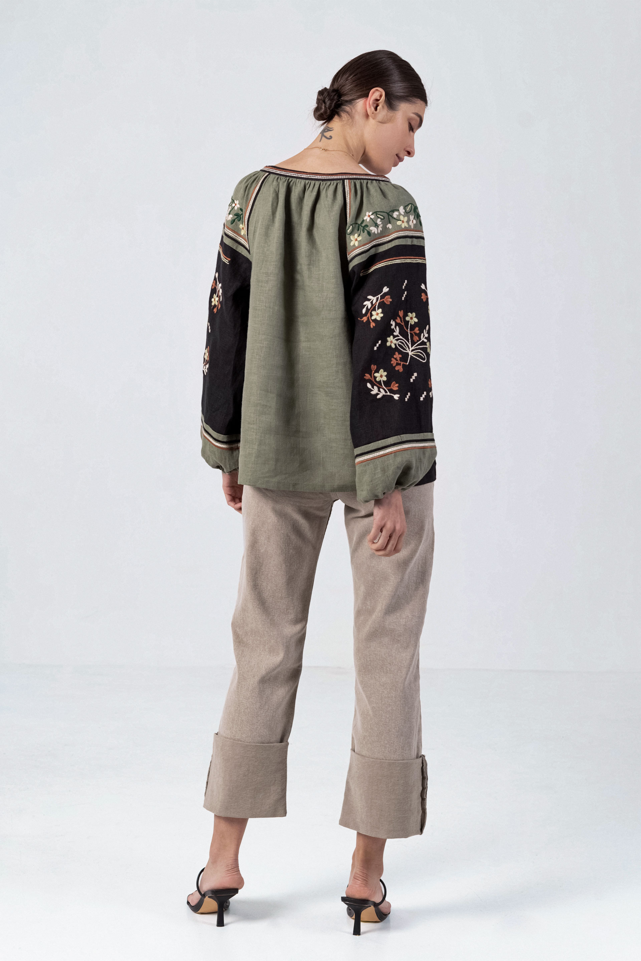 Woman wearing a green and black embroidered blouse with beige pants on a white background