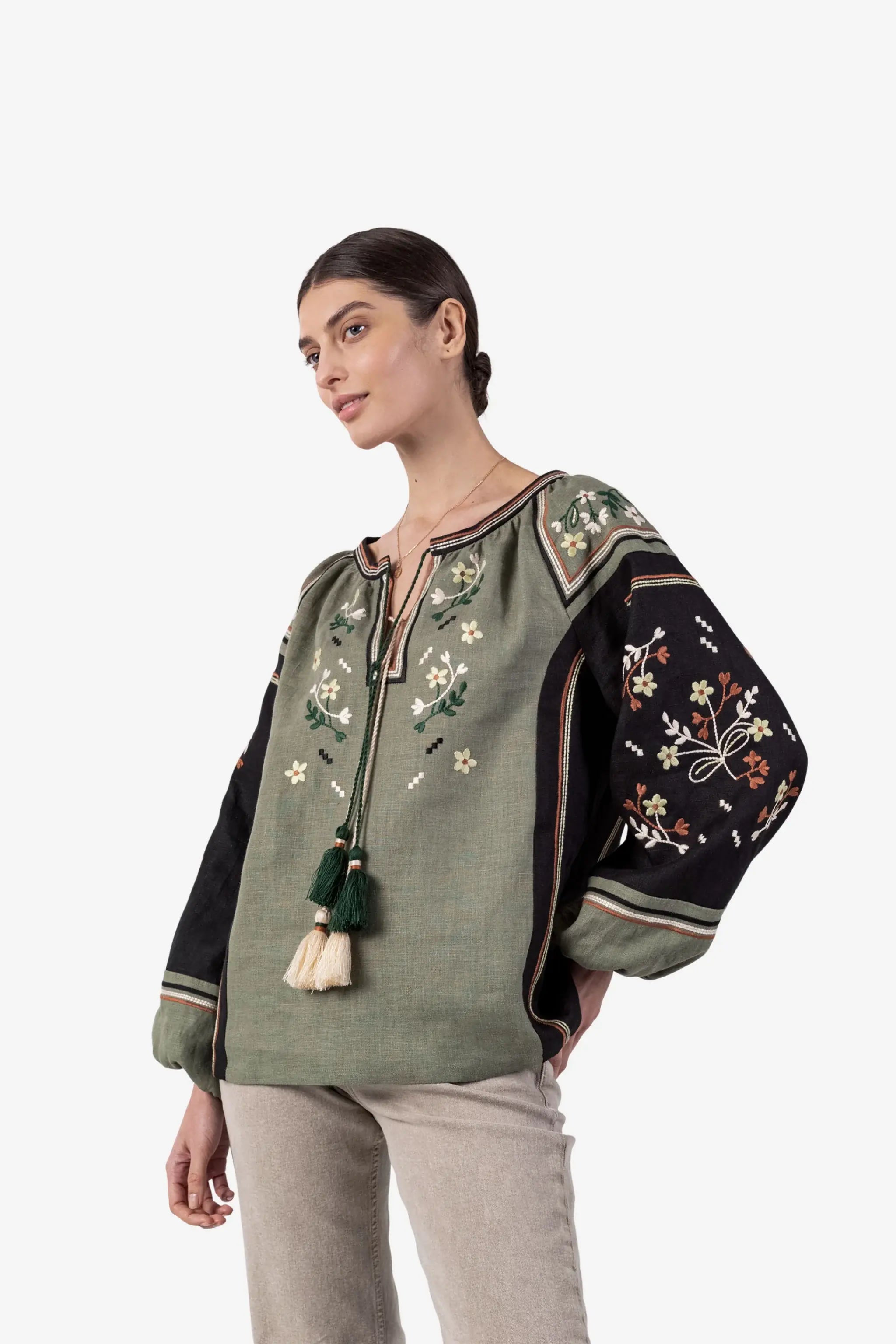 A woman modeling a bohemian-style embroidered blouse with a sage green body, black sleeves, and floral embroidery, paired with light beige trousers, against a plain white background.
