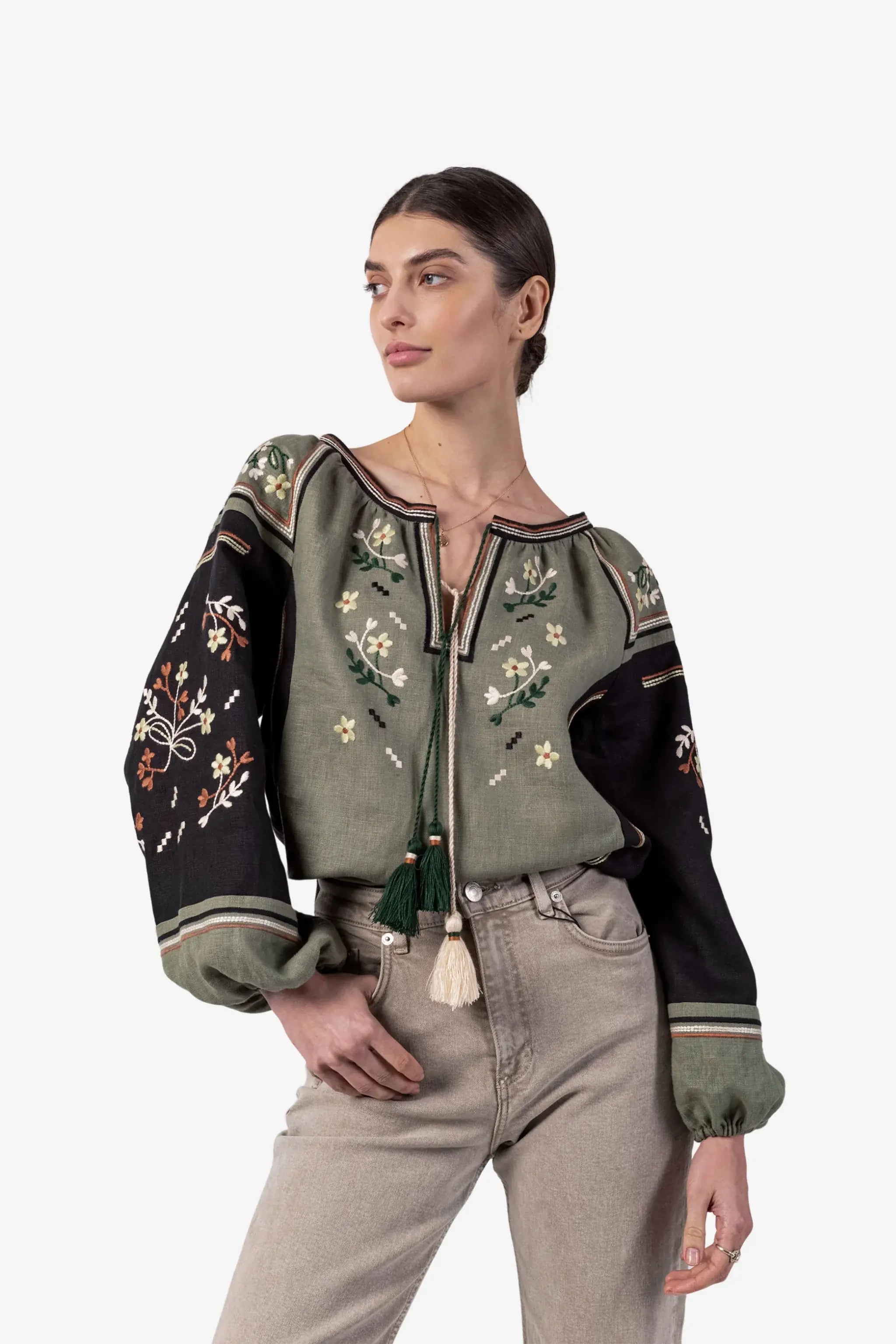 A woman modeling a cropped embroidered blouse with a muted sage green body, black sleeves, and cream floral embroidery, paired with light beige jeans against a plain light gray studio background.