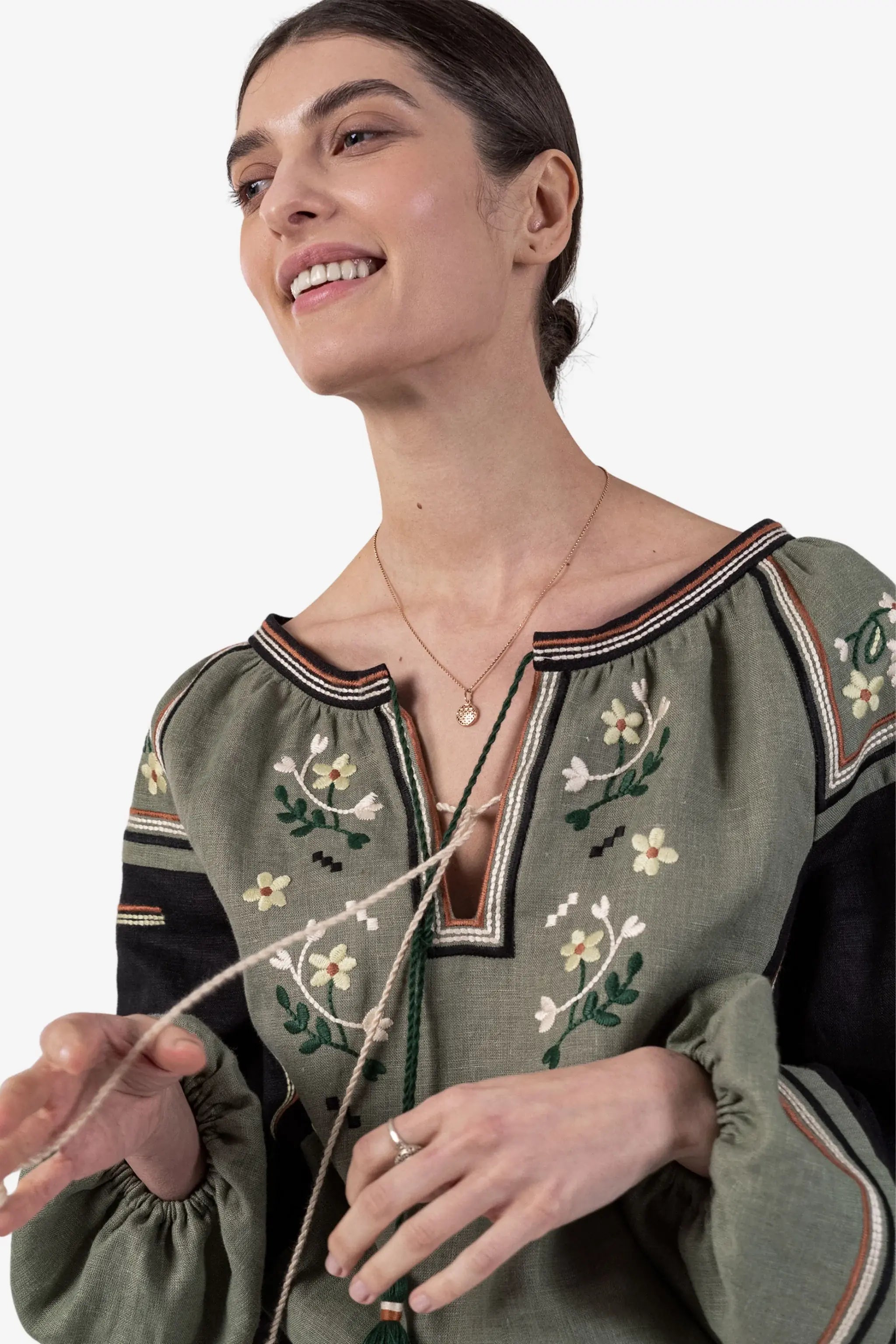 A woman wearing an olive green embroidered blouse with black sleeves and floral embroidery, holding a beaded necklace against a plain white background.