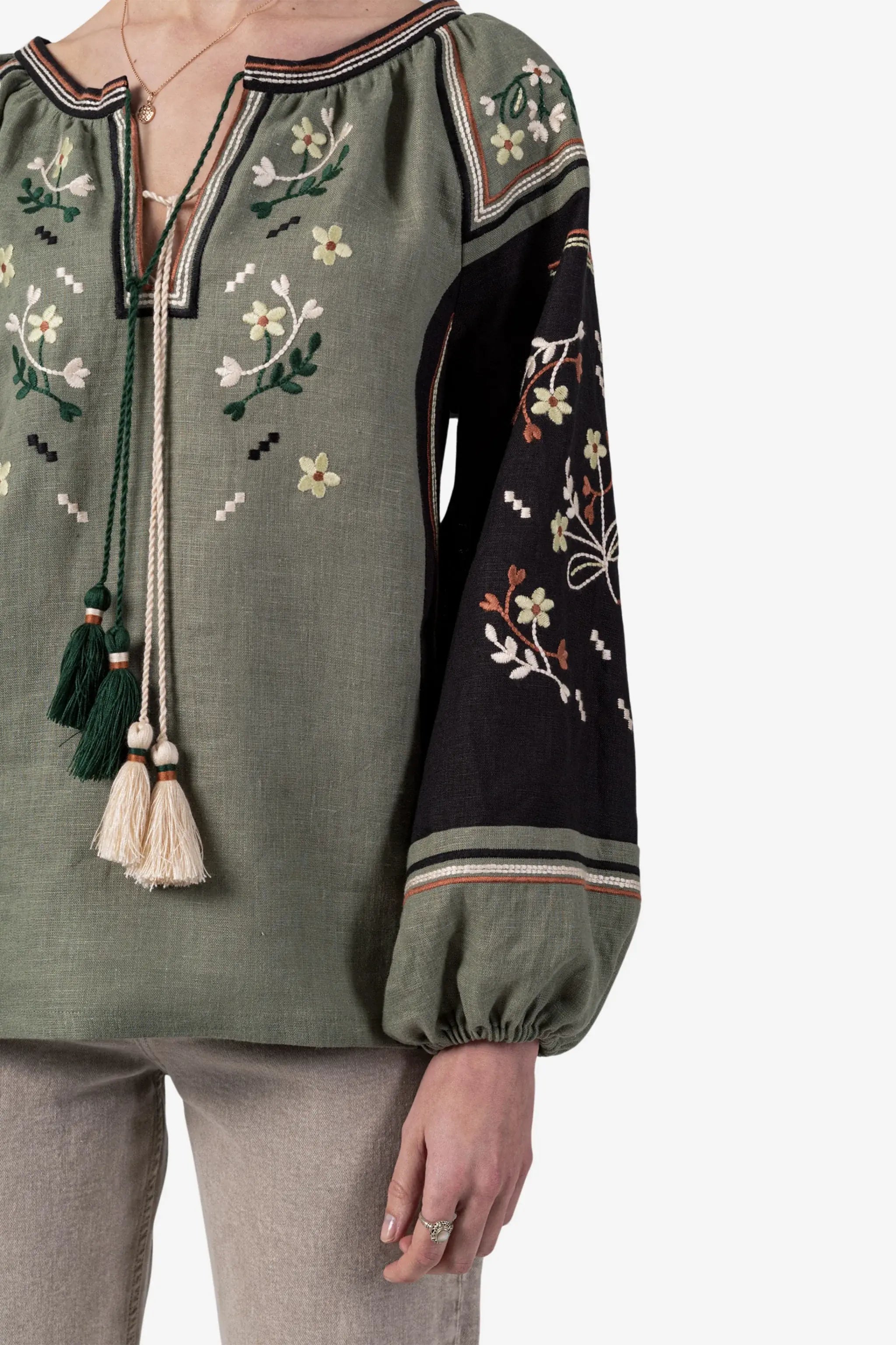 A close-up of a woman wearing a bohemian-style blouse with an olive green body, a black embroidered sleeve, and cream tassels at the neckline, paired with beige trousers against a light gray background.