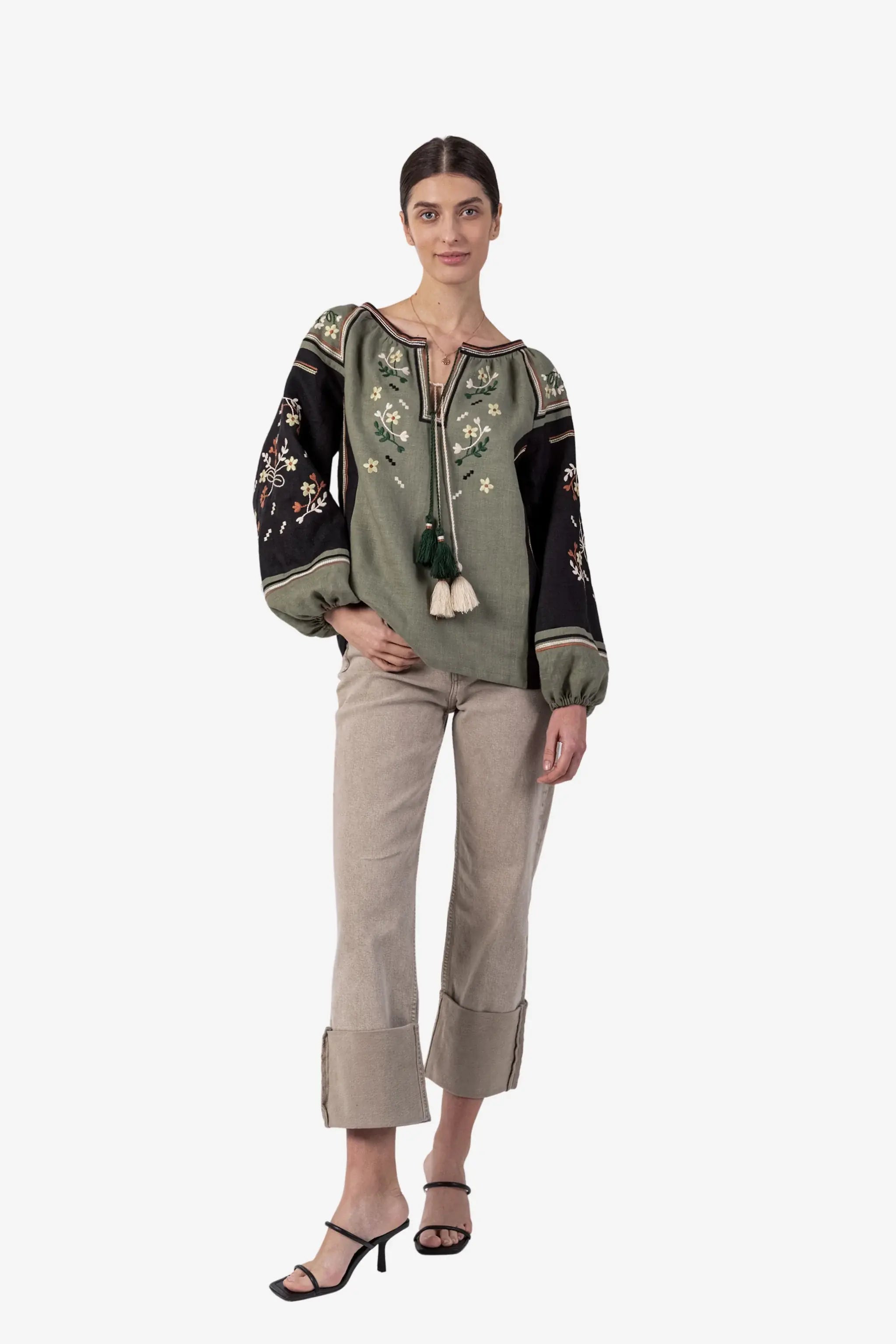 A model stands against a light gray studio background wearing a bohemian-style embroidered blouse in sage green and black with floral patterns, paired with beige rolled-calf trousers and black strappy heels.