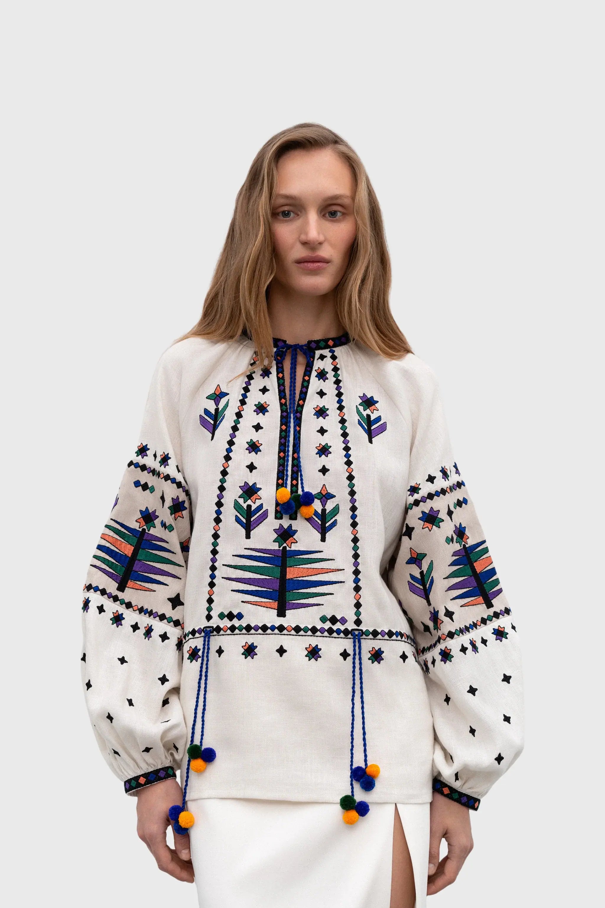 A woman modeling a cream-colored embroidered blouse with colorful geometric patterns and pom-pom tassels, paired with a white skirt, against a light gray studio background.
