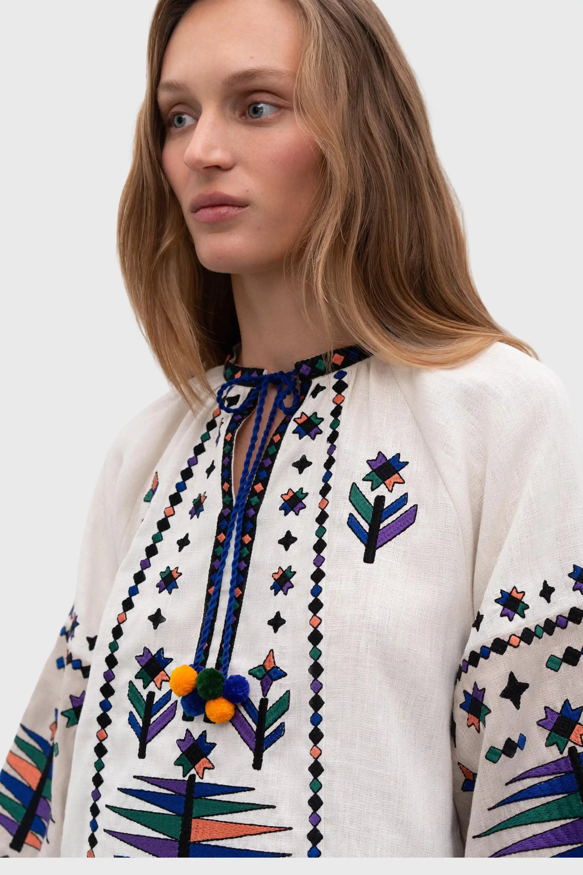 A close-up of a woman wearing a white embroidered blouse with colorful geometric and floral patterns, featuring a tie-neck detail with pom-poms.