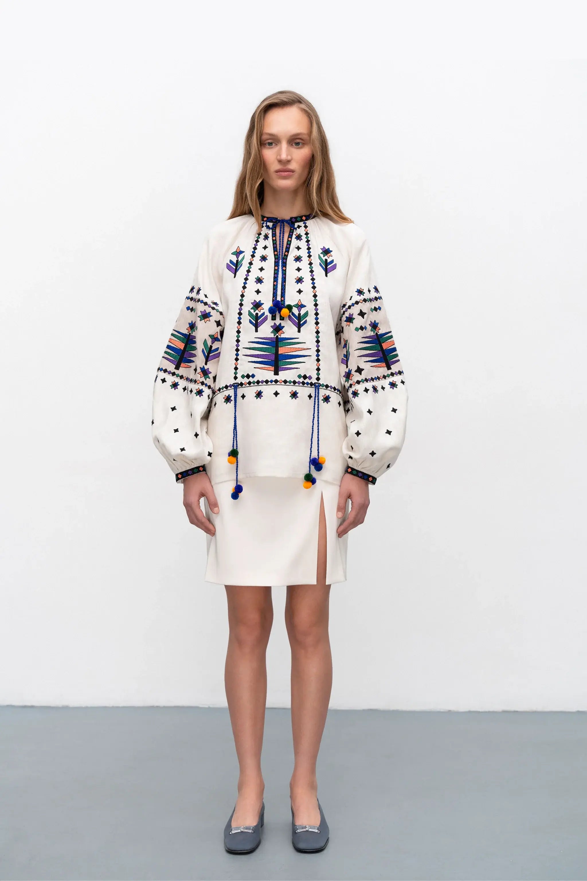 A woman modeling a cream-colored embroidered blouse with blue, yellow, and black geometric patterns, paired with a matching cream skirt and gray flats, set against a plain white studio background.