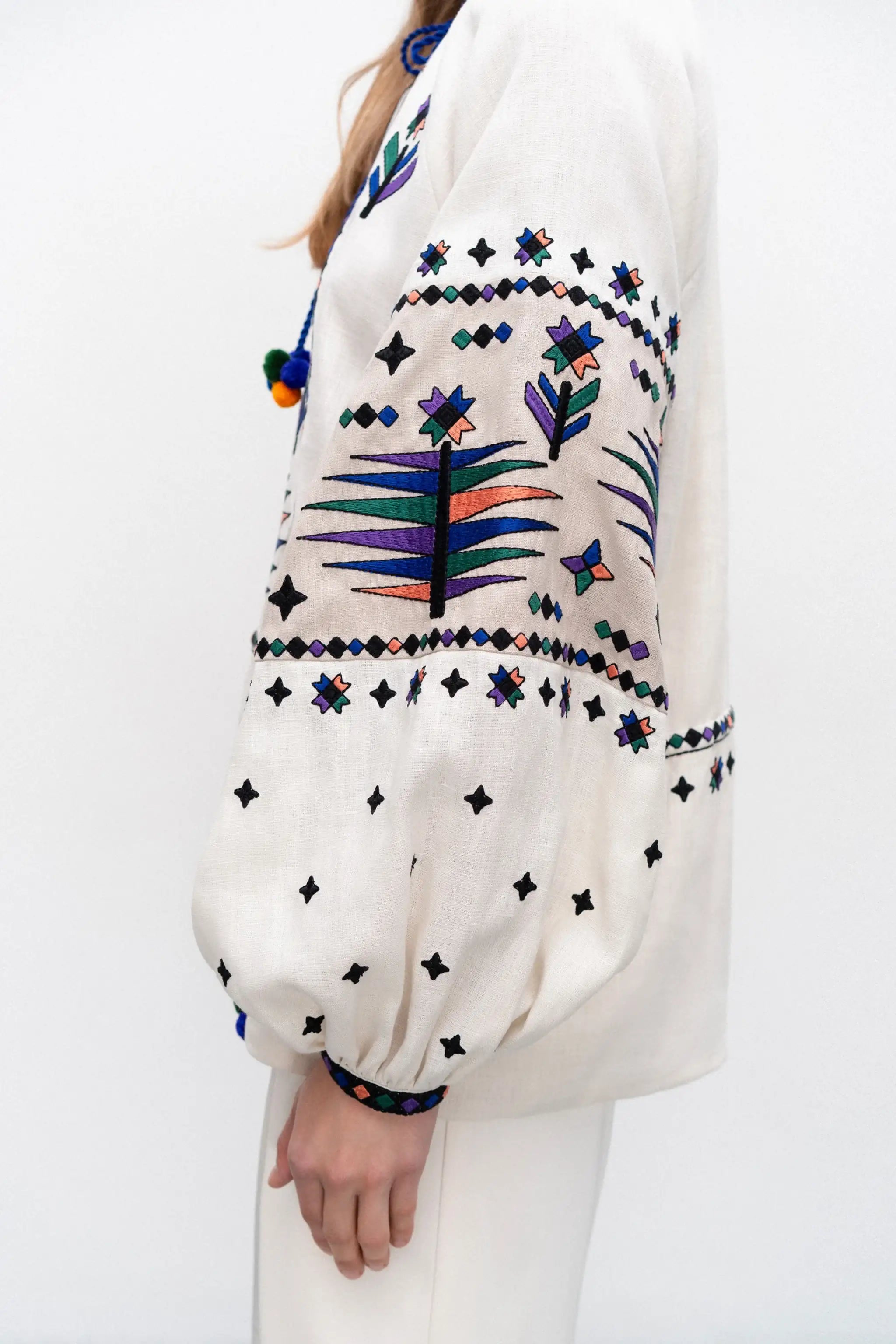 A close-up side view of a person wearing a cream-colored embroidered blouse with colorful geometric and floral patterns, paired with white pants against a plain white background.