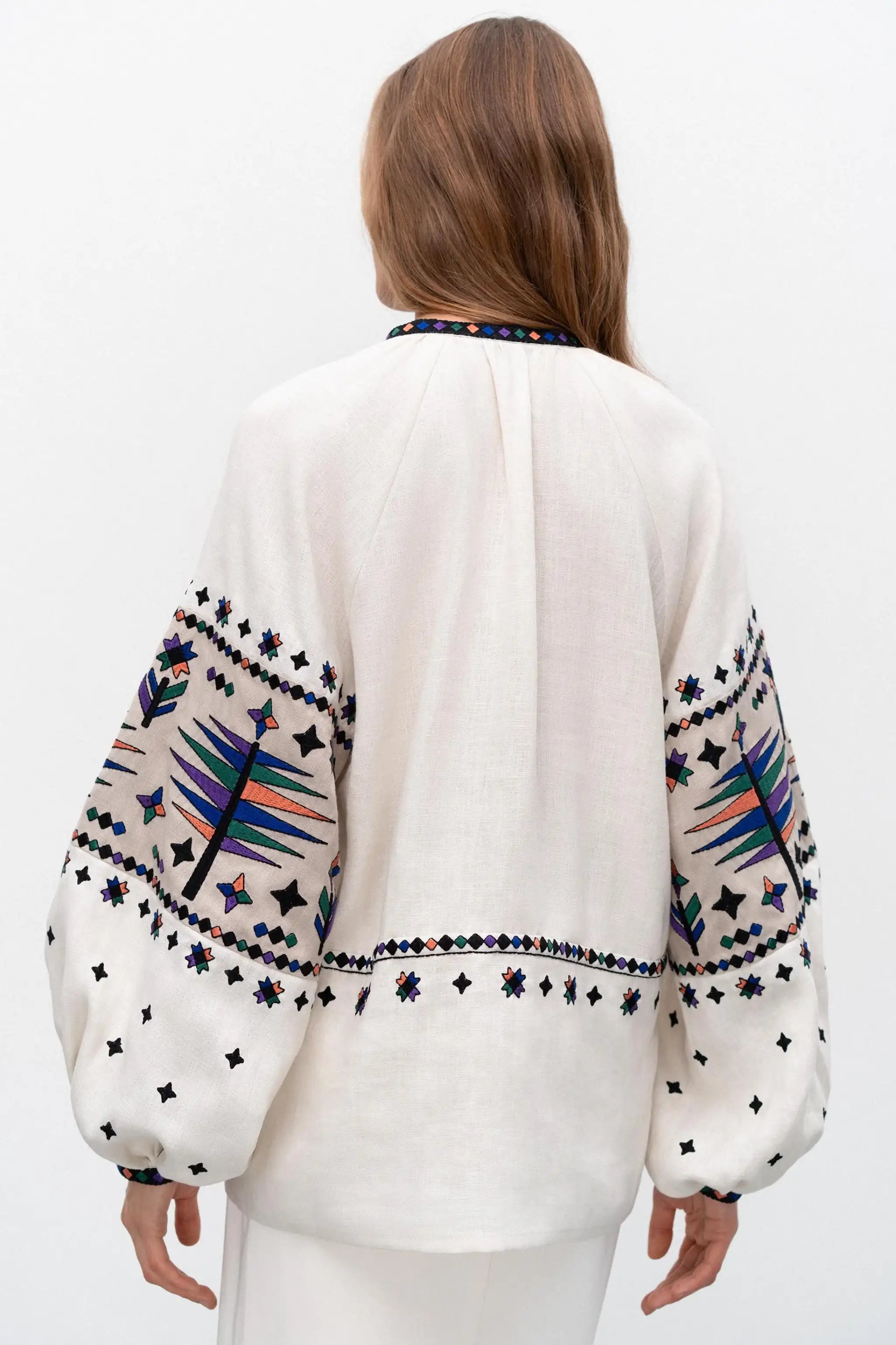 A woman viewed from the back wearing a white embroidered blouse with colorful geometric and star patterns on the sleeves and collar, paired with white pants against a plain white studio background.