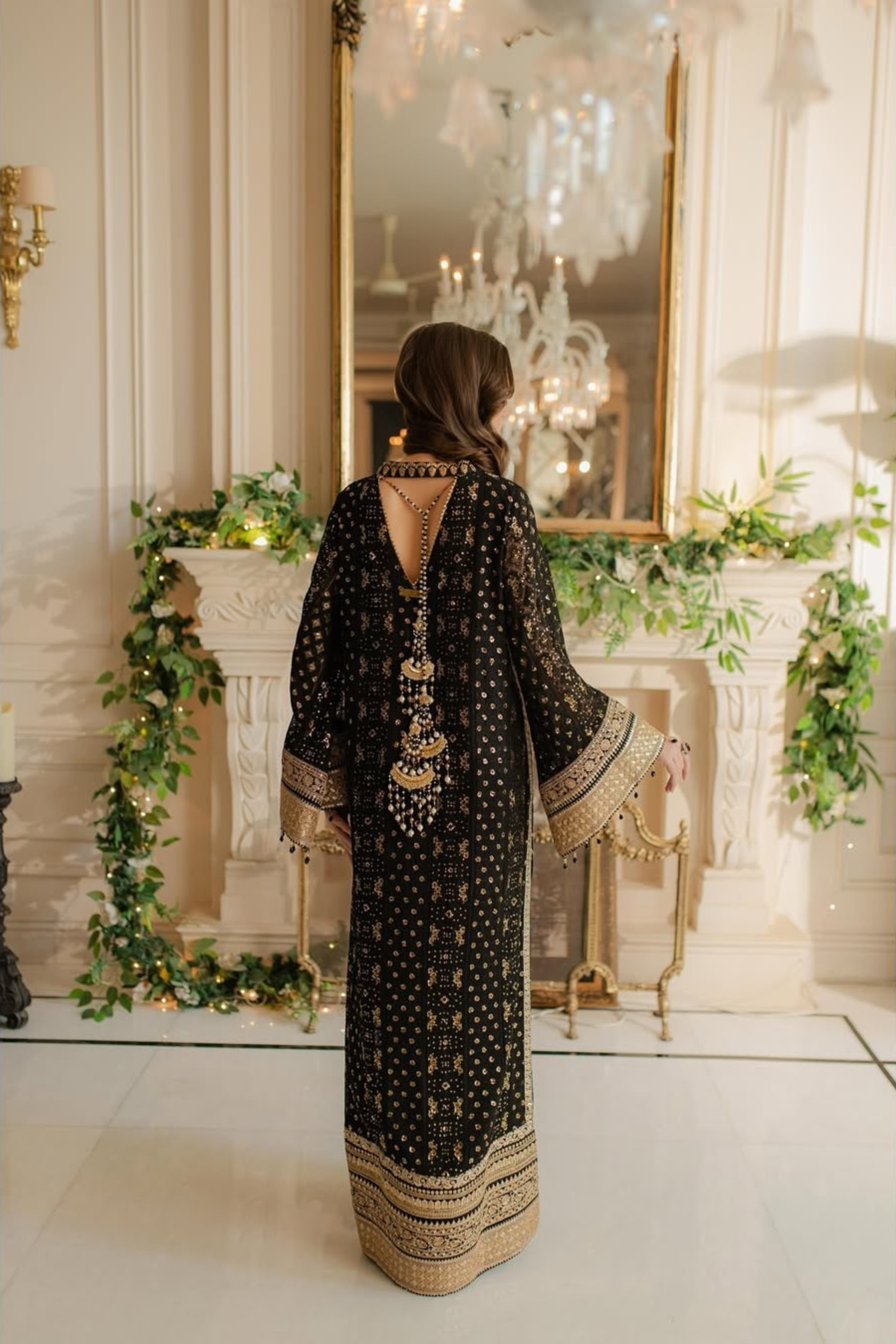 Woman in a black and gold dress standing in a decorated room with a mirror and greenery.