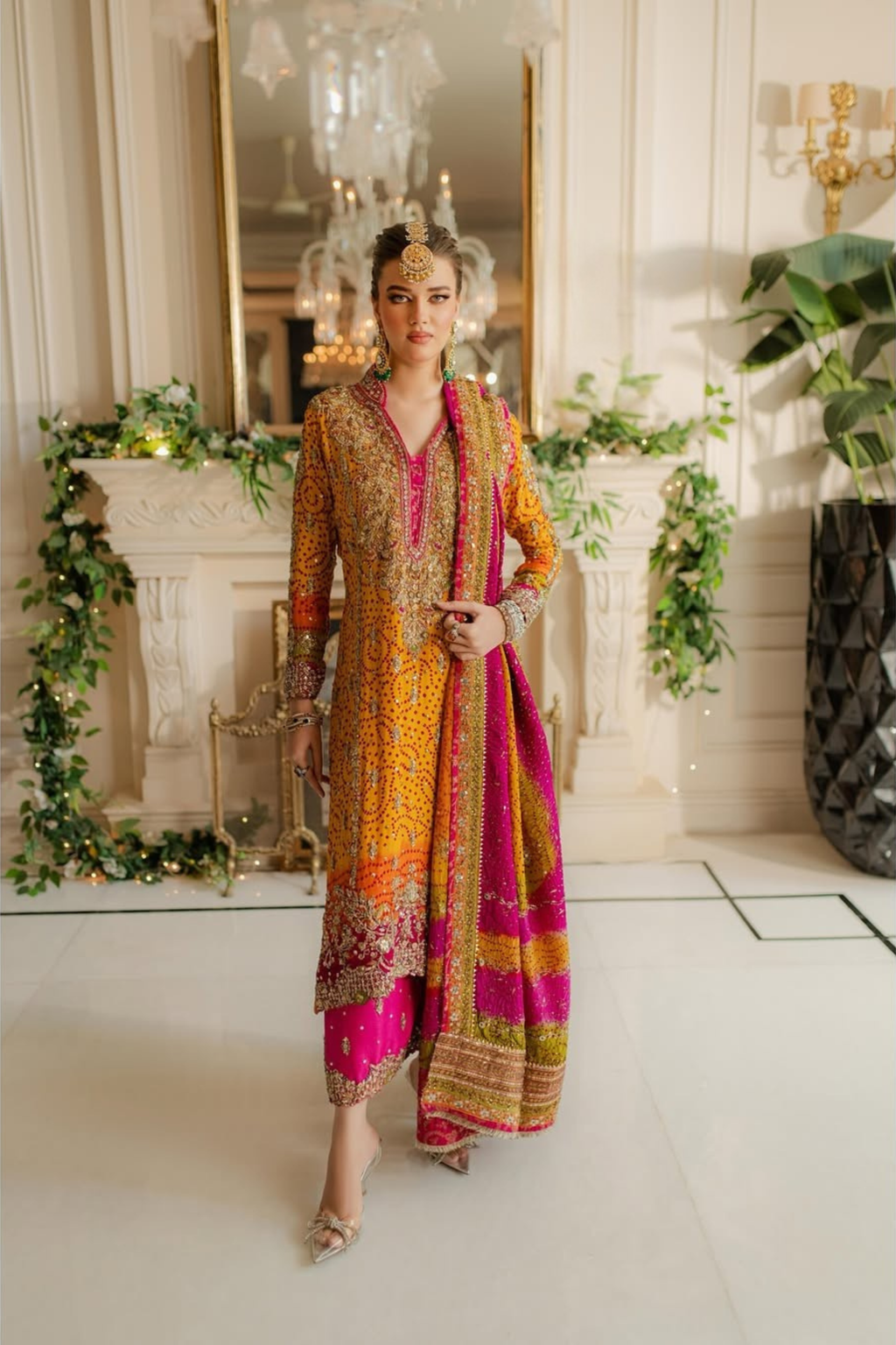 Woman in a colorful traditional outfit standing in a decorated room with greenery and lights.