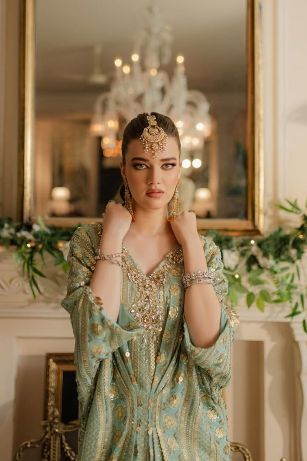 Woman in a green embroidered dress with jewelry in a decorated room with a mirror and chandelier.