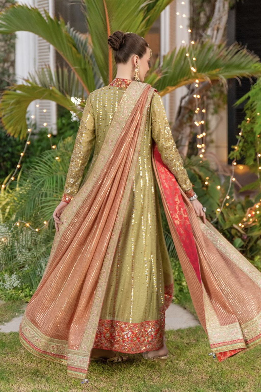 Woman in a traditional green and red outfit with a dupatta, standing outdoors with plants and lights in the background.