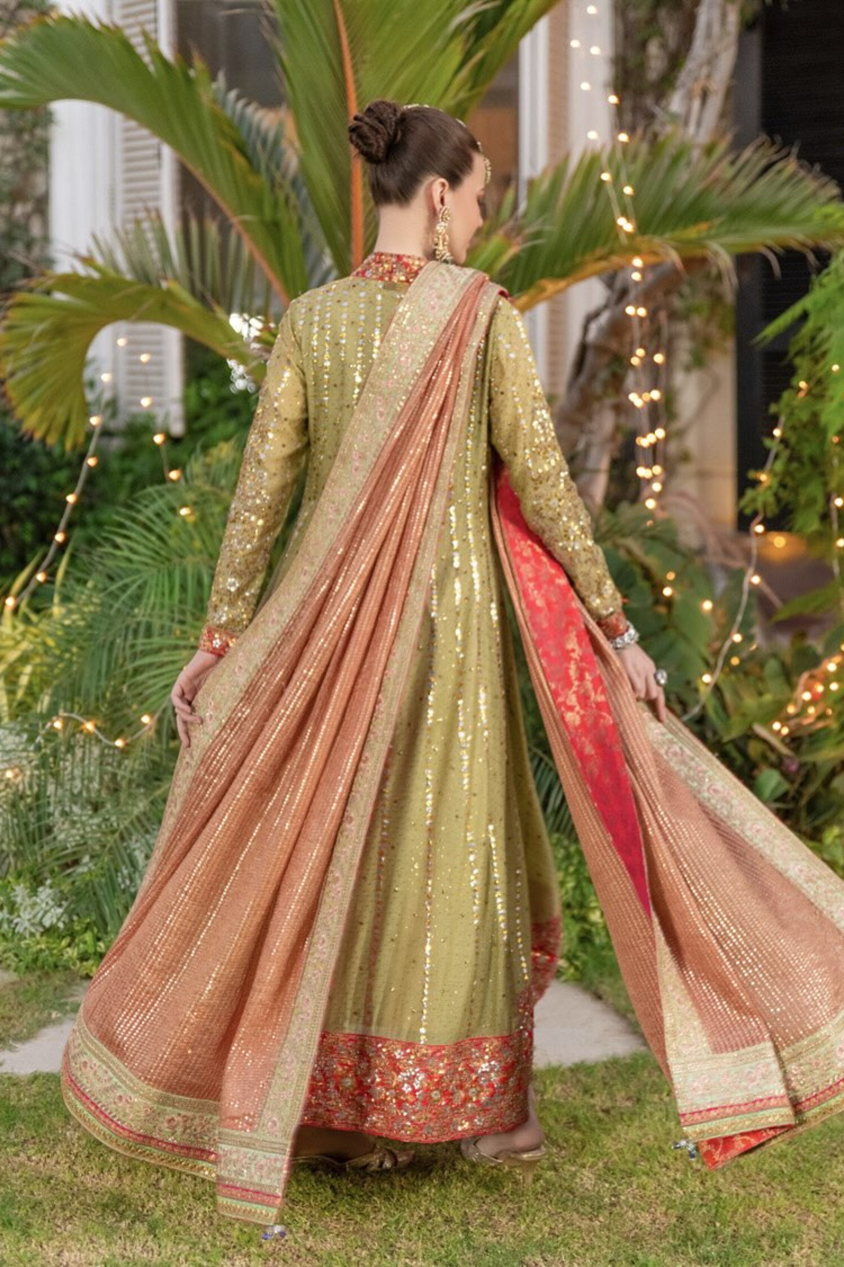 Woman in a traditional green and red outfit with a dupatta, standing outdoors with plants and lights in the background.