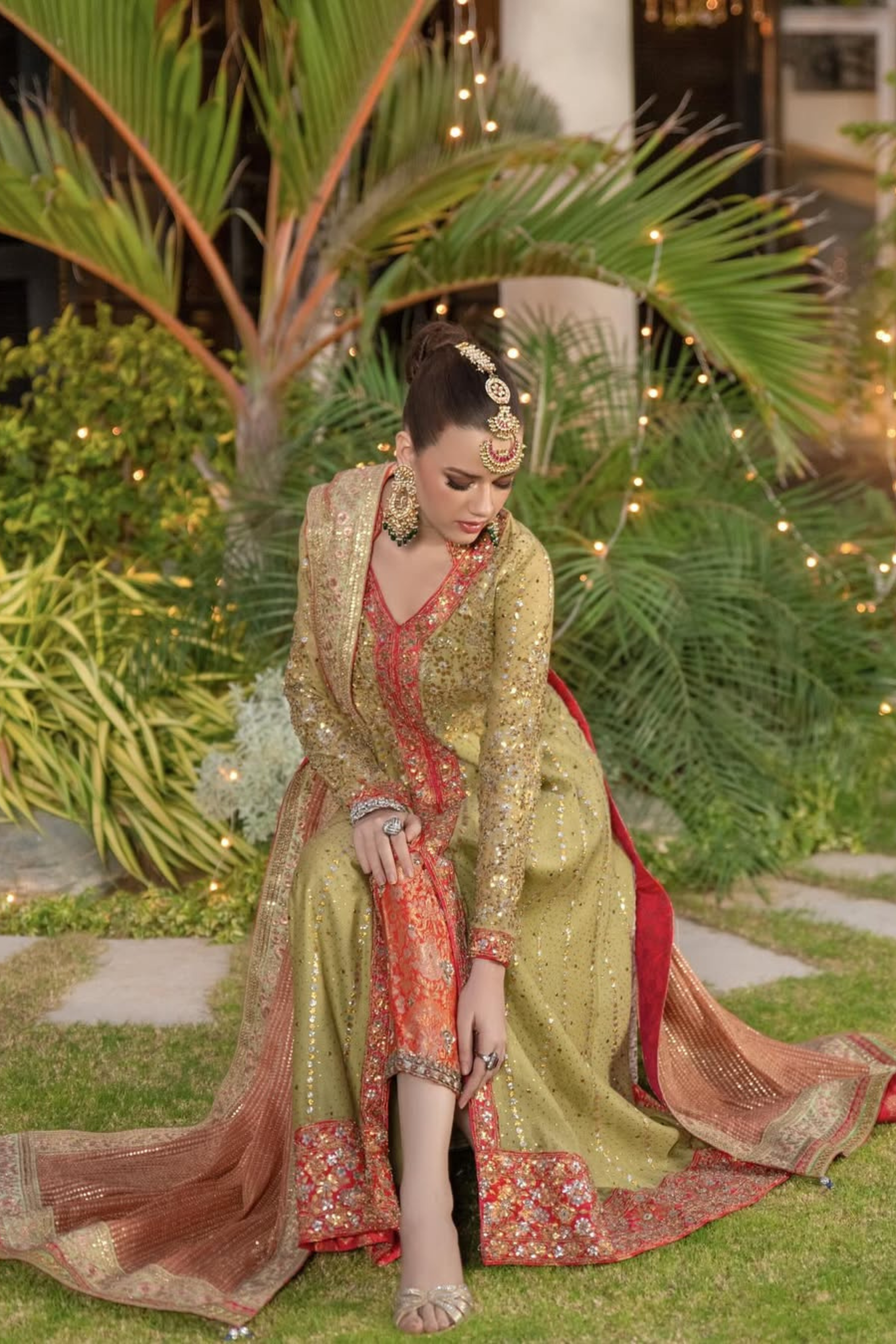 Woman in traditional embroidered outfit sitting outdoors with greenery and lights in the background