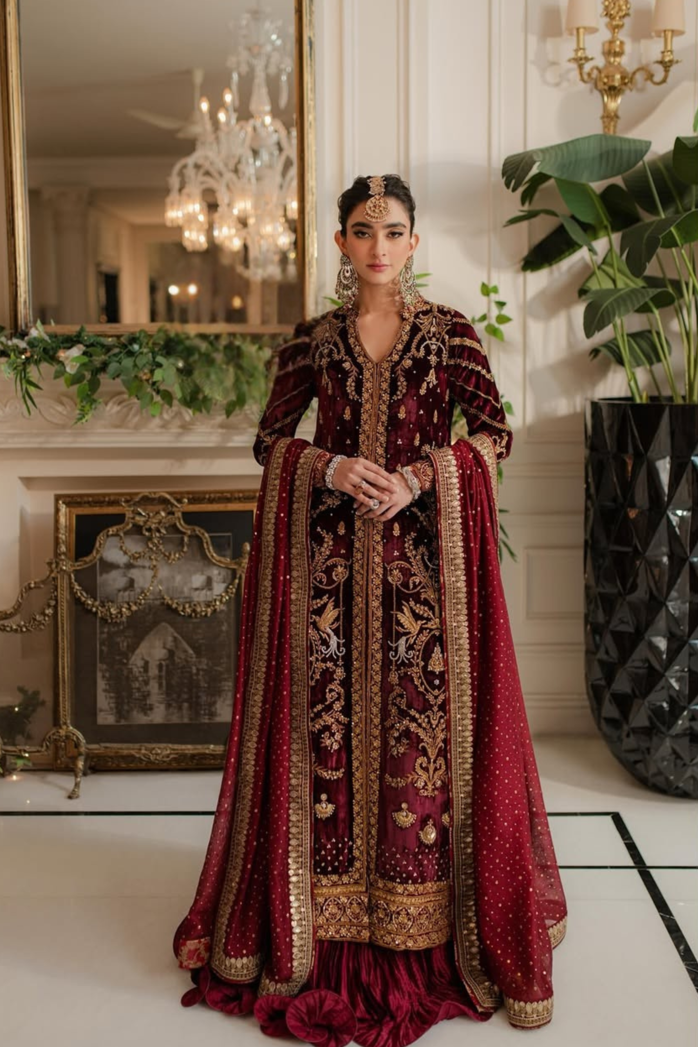 Woman in a red and gold traditional outfit standing in an elegant room with decorative elements.