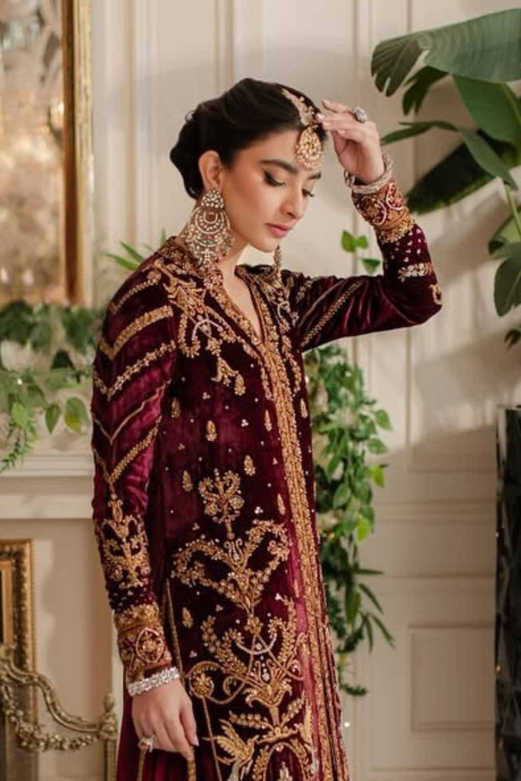 Woman in a maroon and gold embroidered traditional outfit with jewelry, standing indoors.