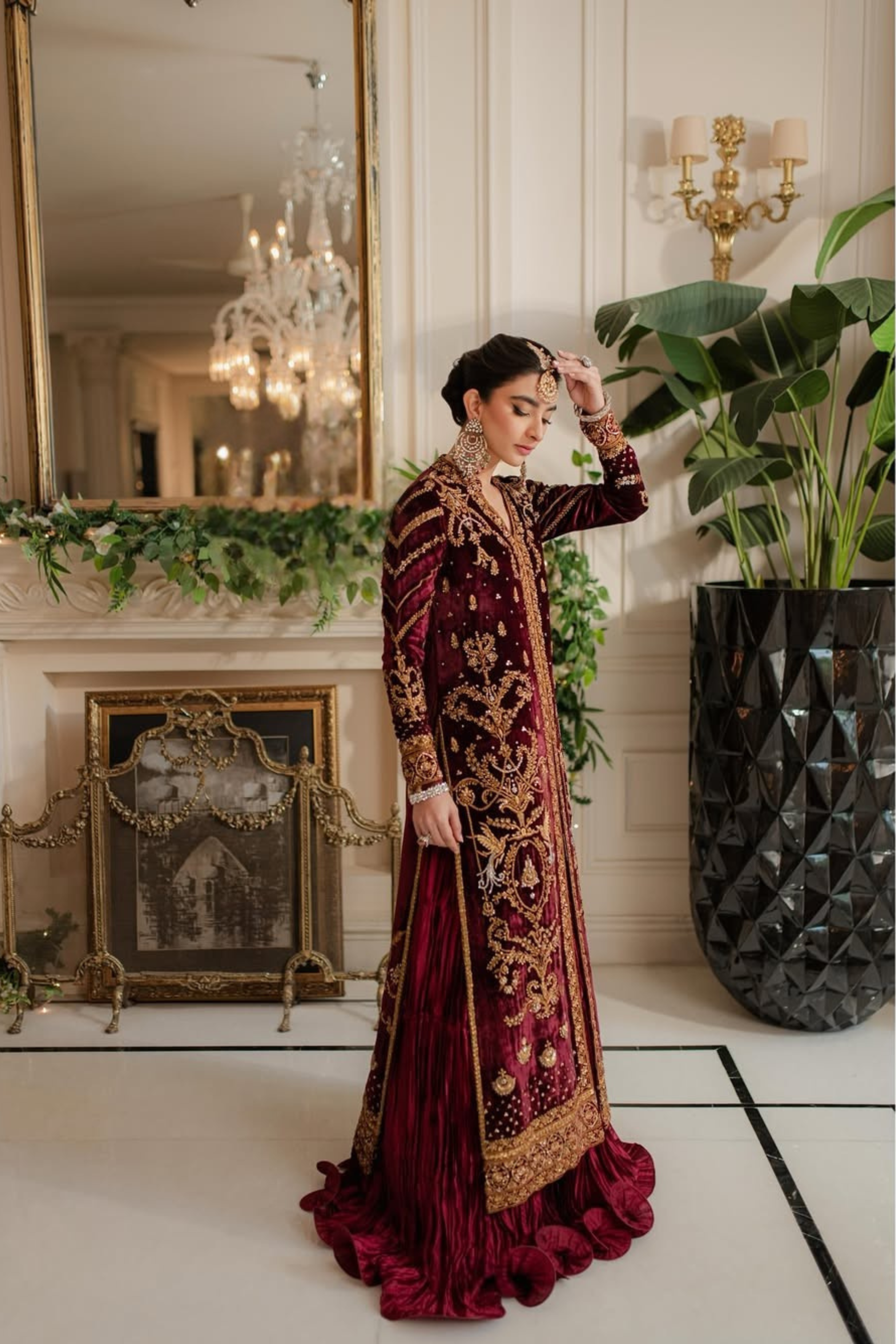 Woman in a red and gold embroidered traditional outfit standing in an elegant room with chandelier and fireplace.