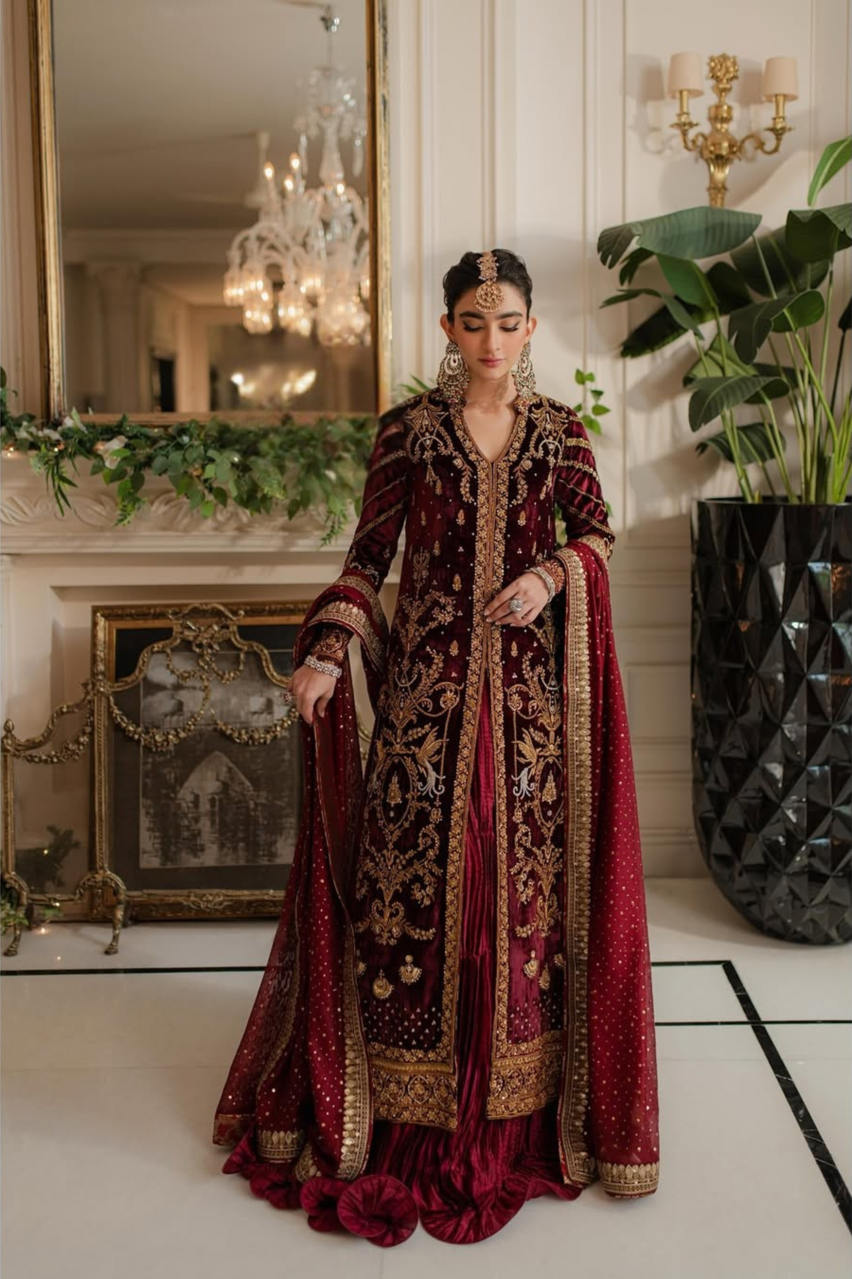 Woman in an ornate red and gold dress standing in a decorated room with a mirror and plants.