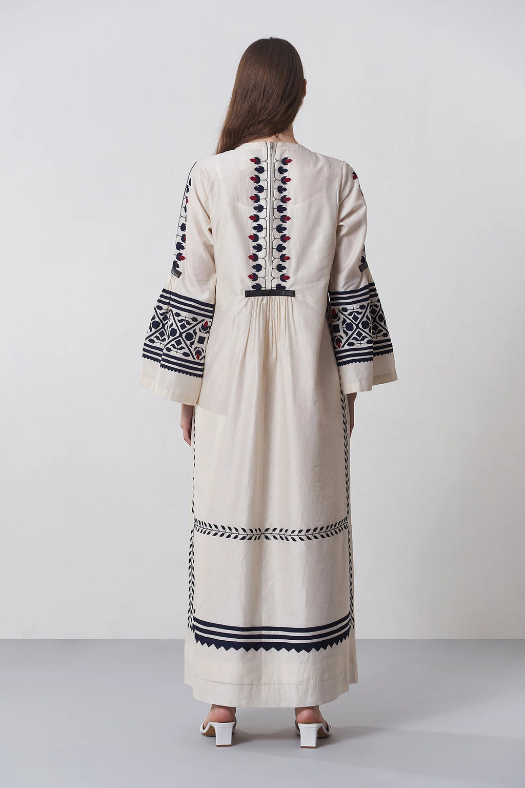 an russian model wearing An ivory floor-length Chanderi dress featuring dramatic bell sleeves and vertical panels of tonal geometric hand-cut appliqué.
