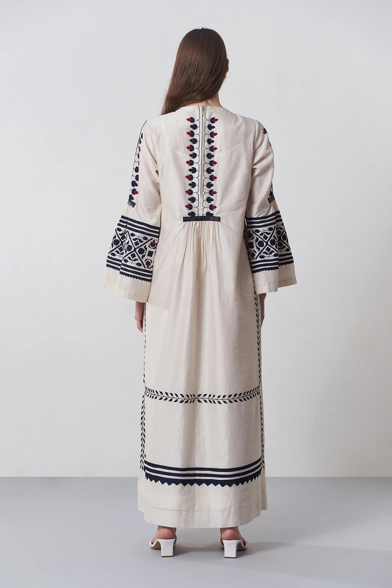 an russian model wearing An ivory floor-length Chanderi dress featuring dramatic bell sleeves and vertical panels of tonal geometric hand-cut appliqué.