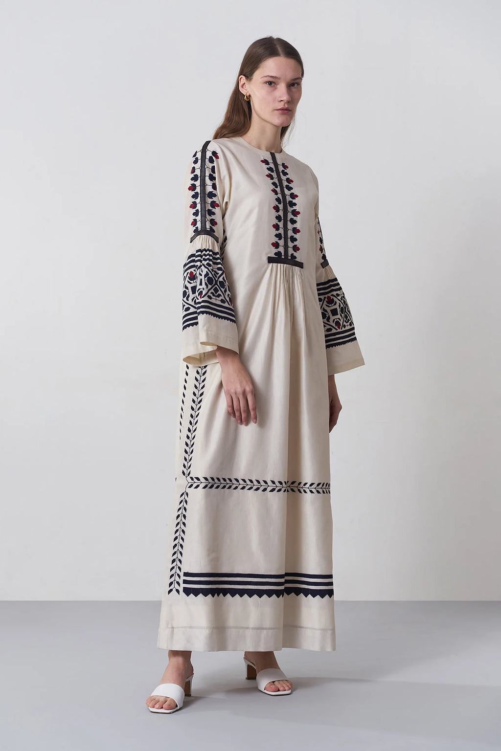 an russian model wearing An ivory floor-length Chanderi dress featuring dramatic bell sleeves and vertical panels of tonal geometric hand-cut appliqué.