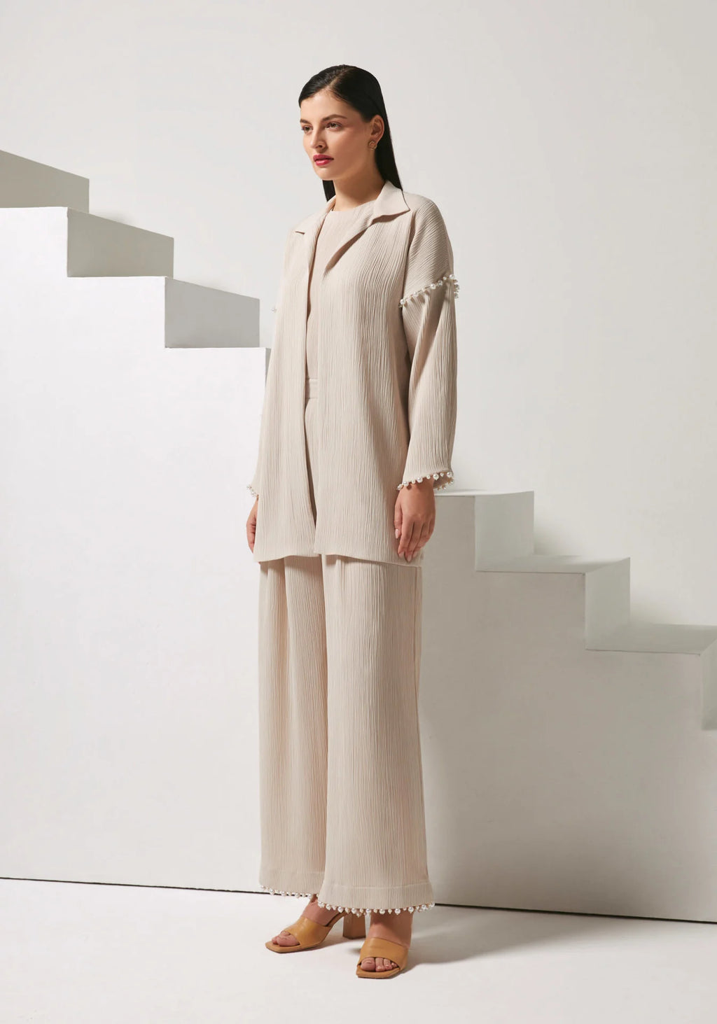 A woman models a light beige two-piece linen set consisting of a long-sleeve button-up blazer with decorative pom-pom trim on the cuffs and matching wide-leg trousers, posed in front of minimalist white geometric stairs.