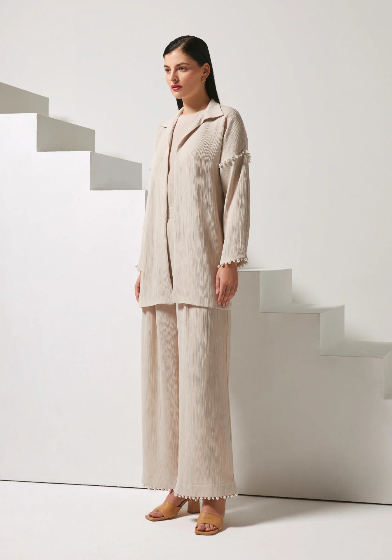 A woman models a light beige two-piece linen set consisting of a long-sleeve button-up blazer with decorative pom-pom trim on the cuffs and matching wide-leg trousers, posed in front of minimalist white geometric stairs.