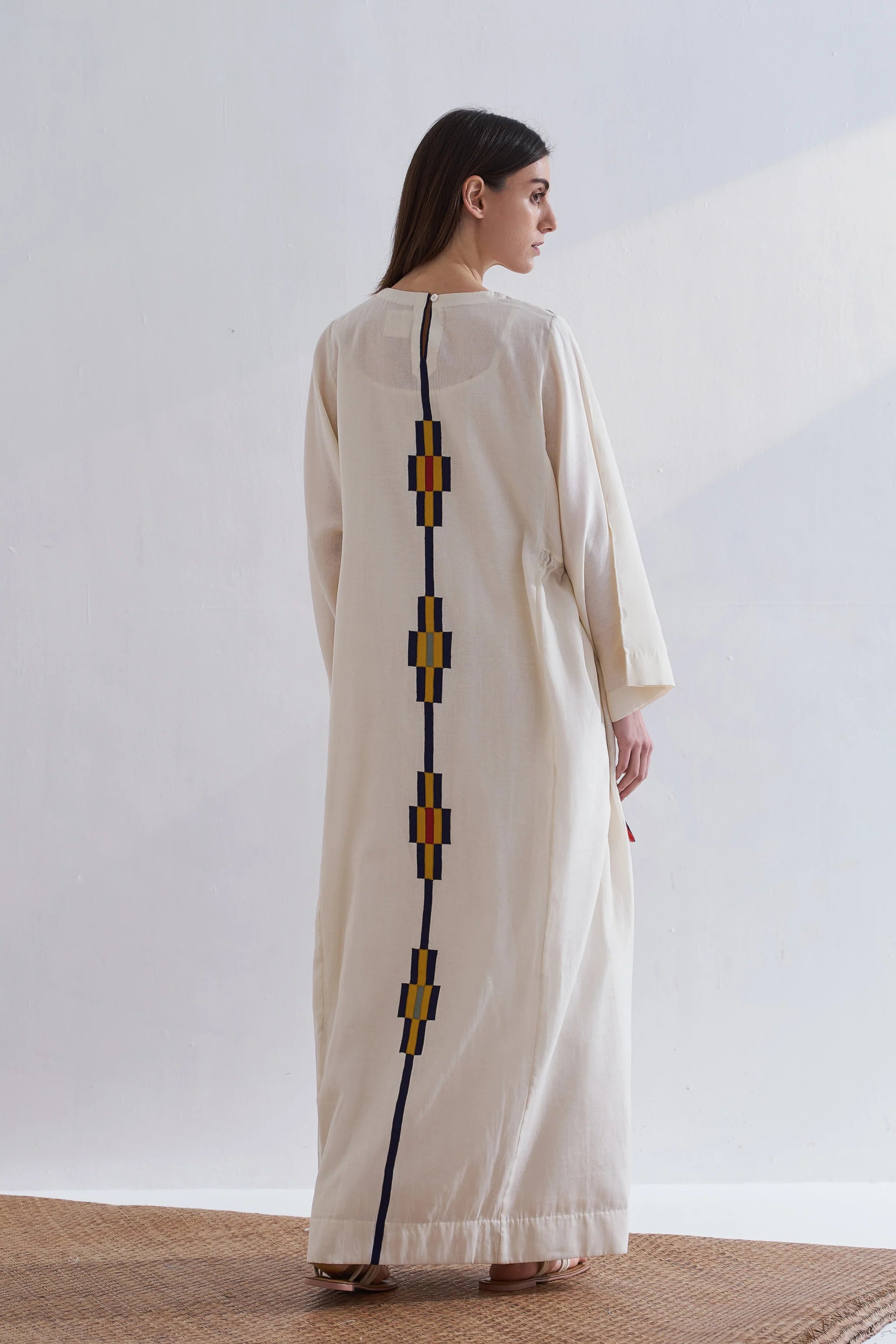 Woman wearing a long, light-colored dress with geometric patterns on a plain background