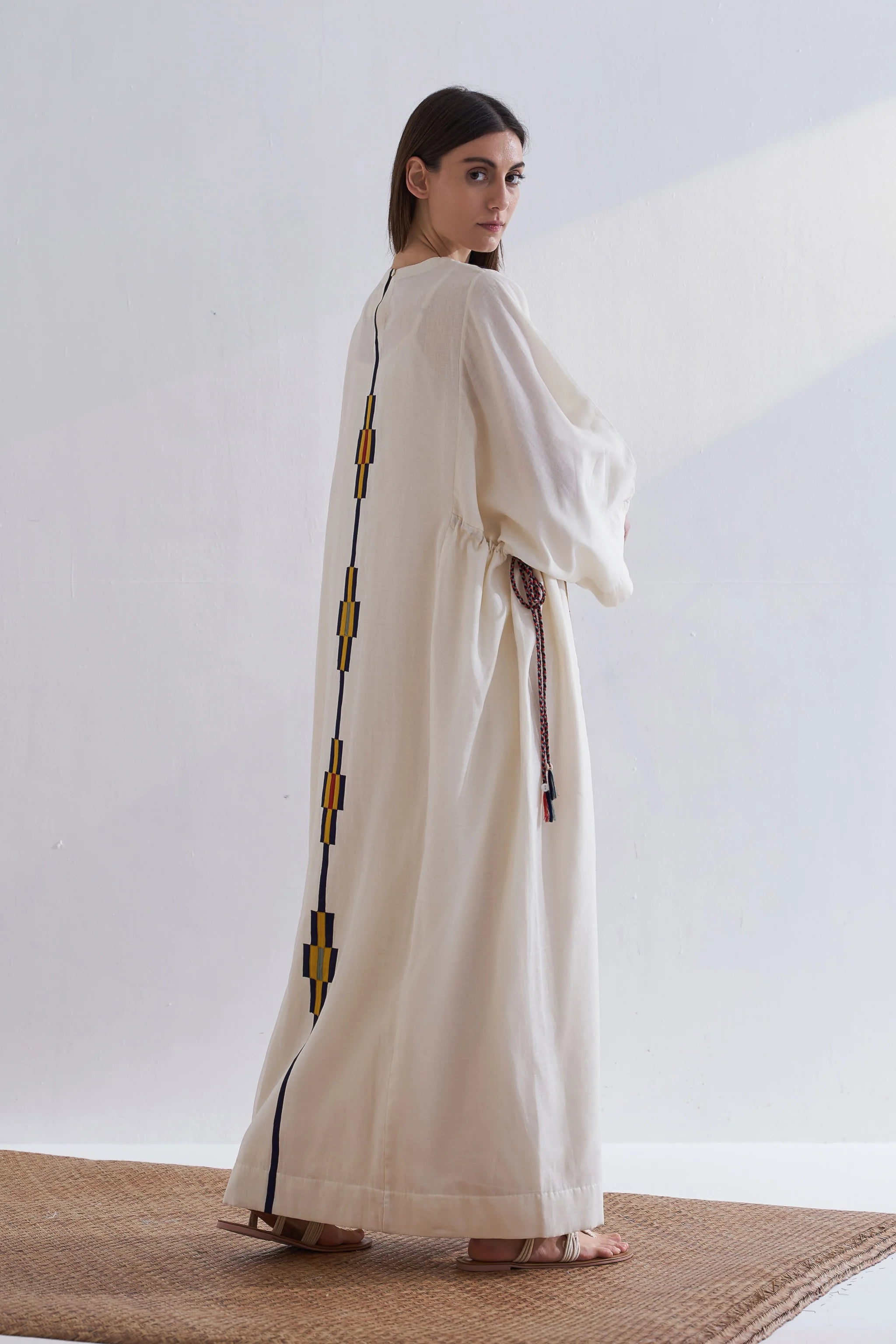 Woman wearing a long, light-colored dress with decorative elements on a plain background