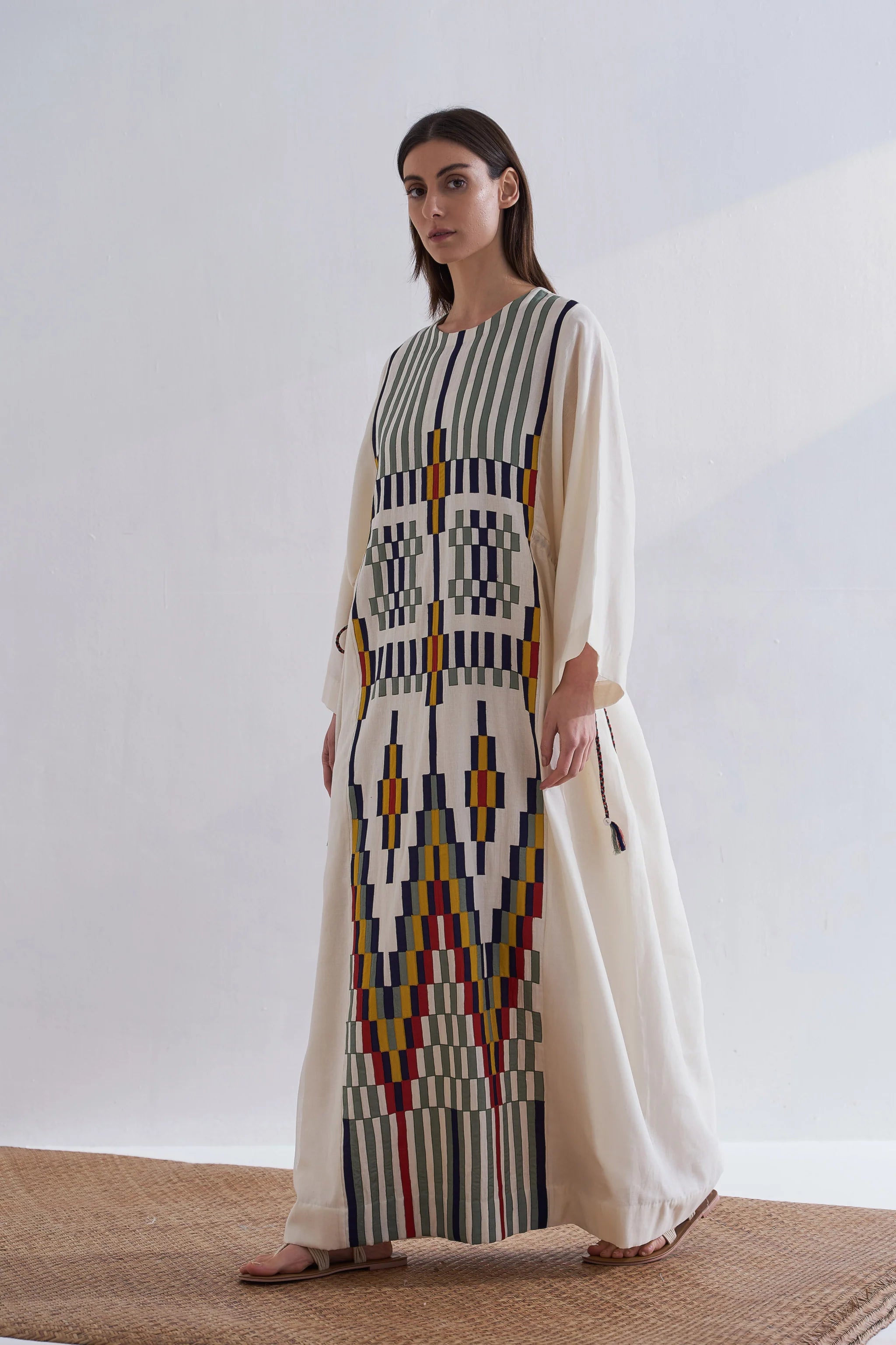 A model in a cream Cotton Silk Chanderi kaftan dress with vertical green stripes and a complex geometric multi-colored appliqué panel on the front and back.