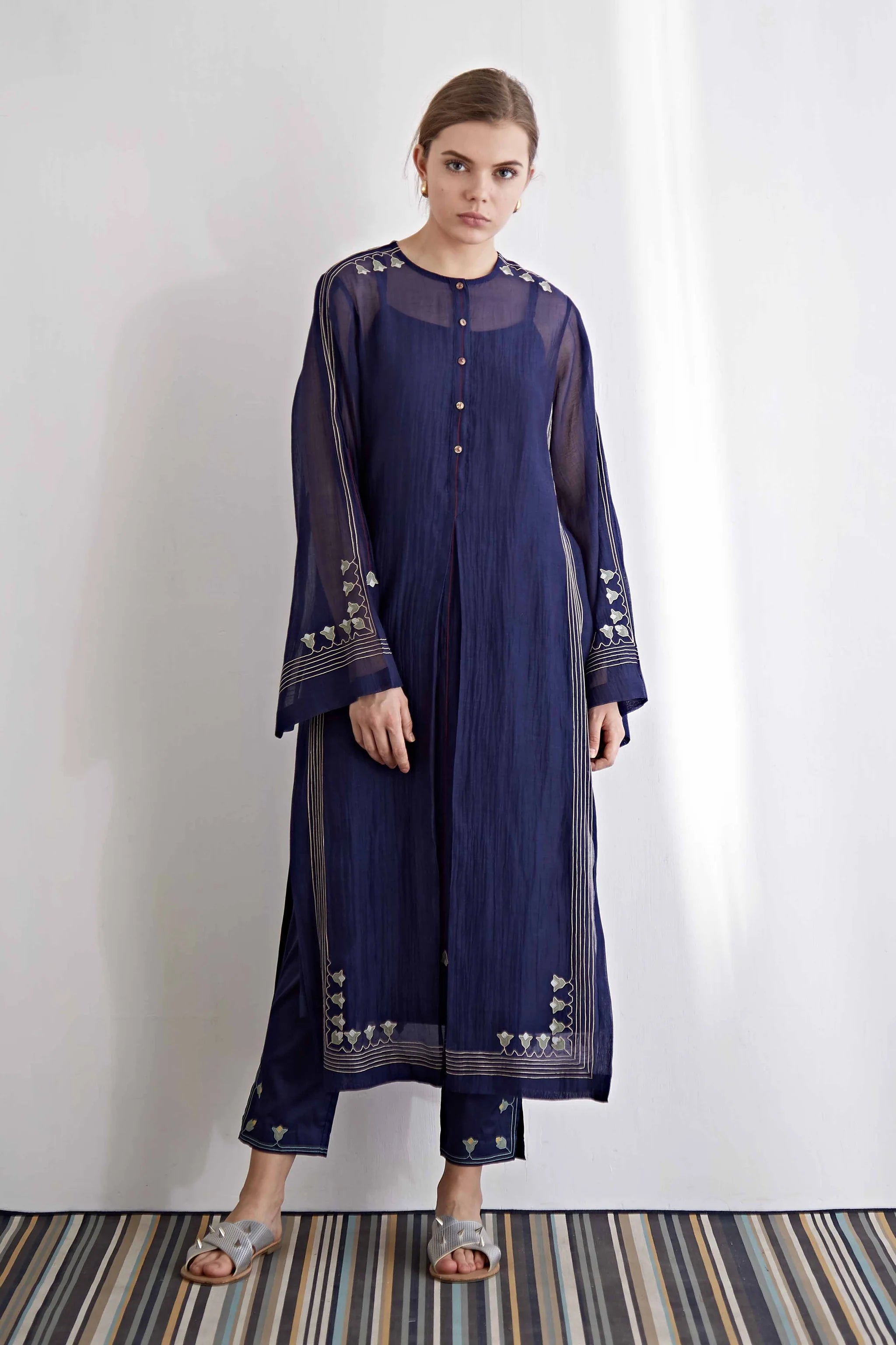 A model in a navy blue long kameez with center pleat detailing and white floral hand-cut appliqué along the hem and sleeves.
