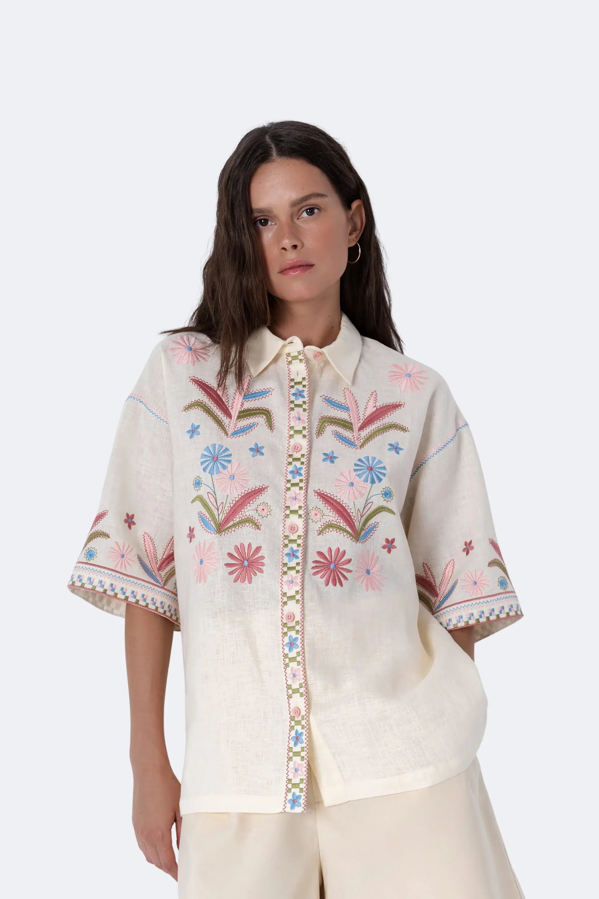 A woman modeling a cream-colored short-sleeve button-up shirt with colorful floral embroidery on the front and sleeves, paired with matching cream shorts and brown slide sandals, against a light gray studio background.