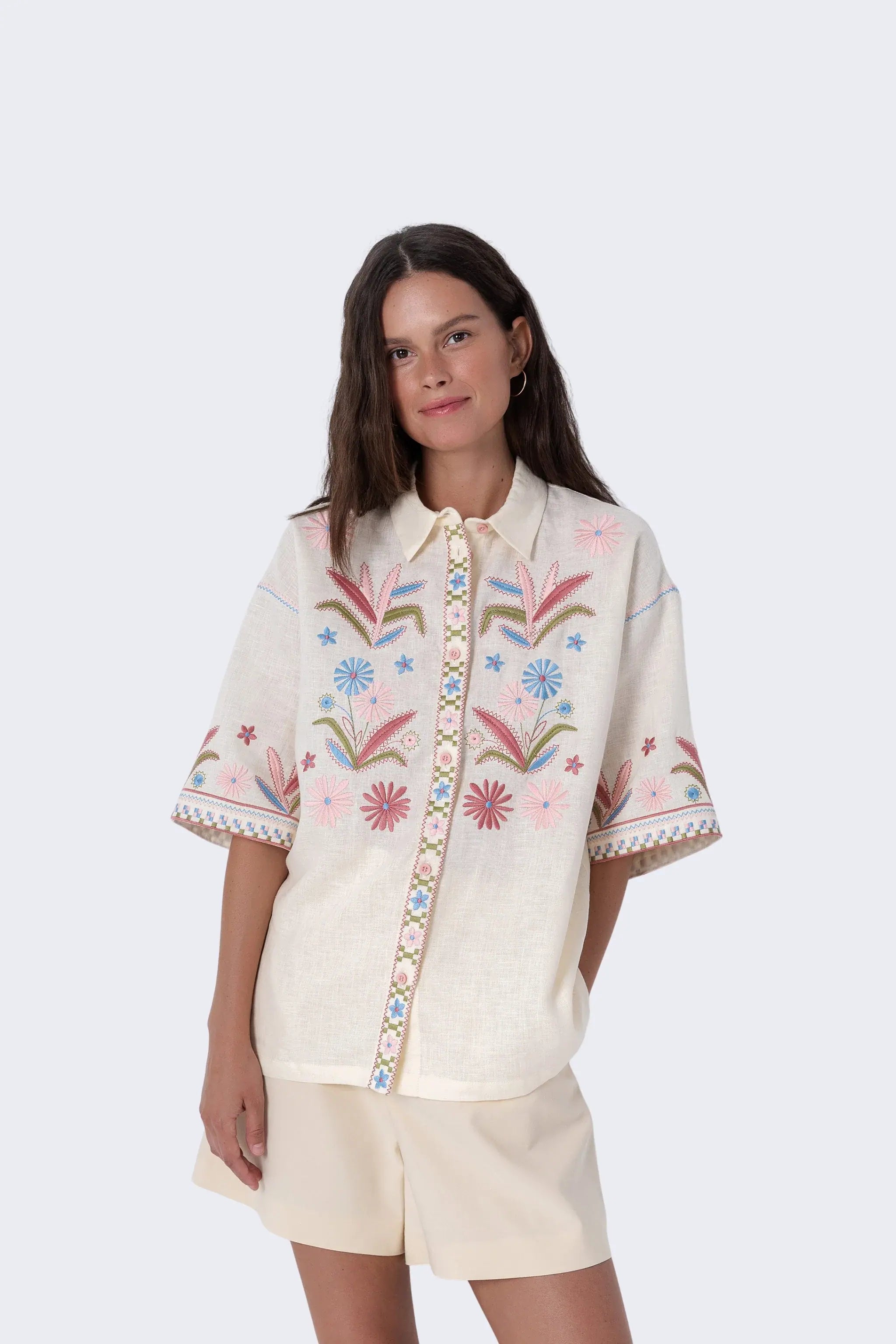 A woman modeling a cream-colored short-sleeve button-up shirt with colorful floral embroidery on the front and sleeves, paired with matching cream shorts and brown slide sandals, against a light gray studio background.