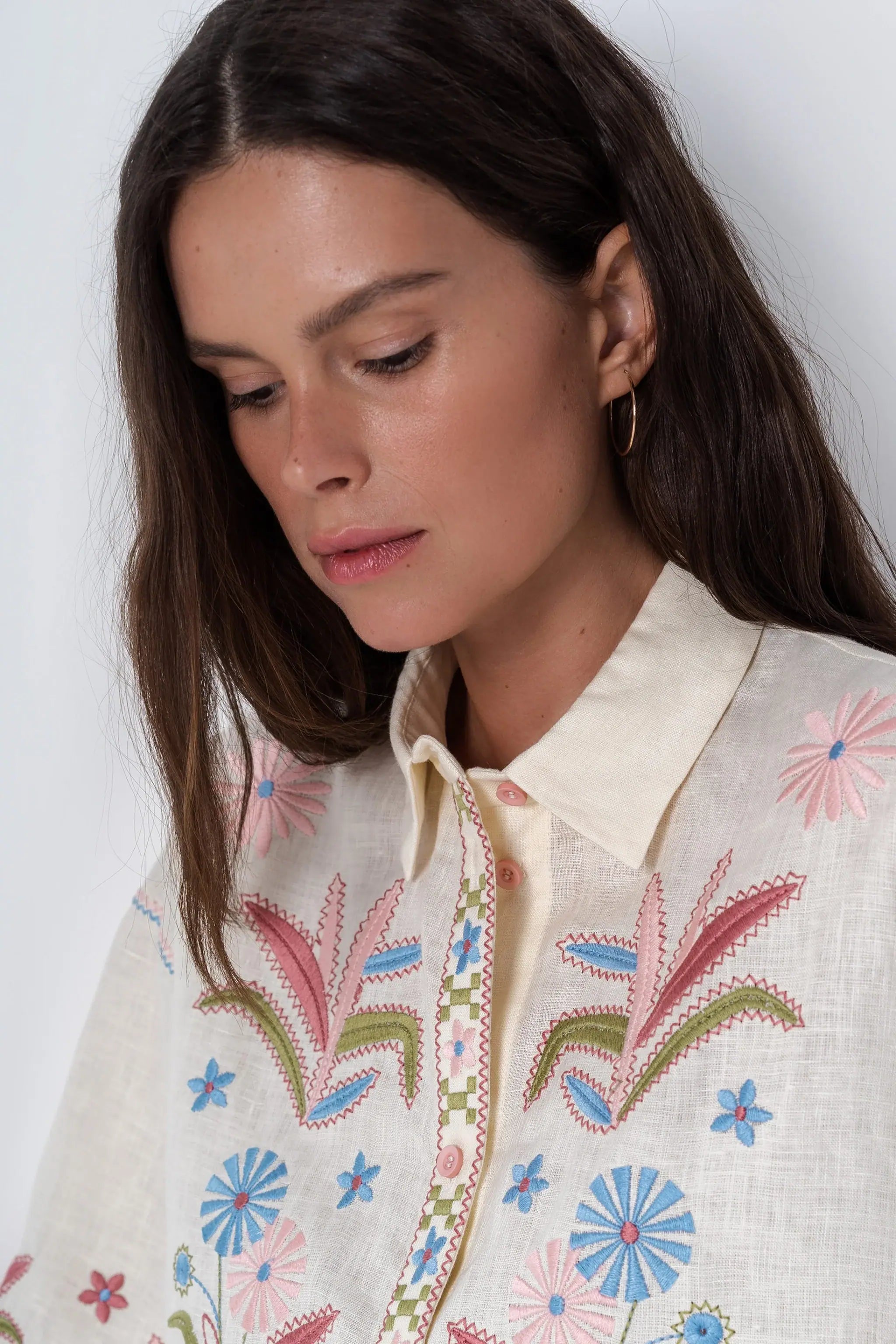 A woman modeling a cream-colored short-sleeve button-up shirt with colorful floral embroidery on the front and sleeves, paired with matching cream shorts and brown slide sandals, against a light gray studio background.