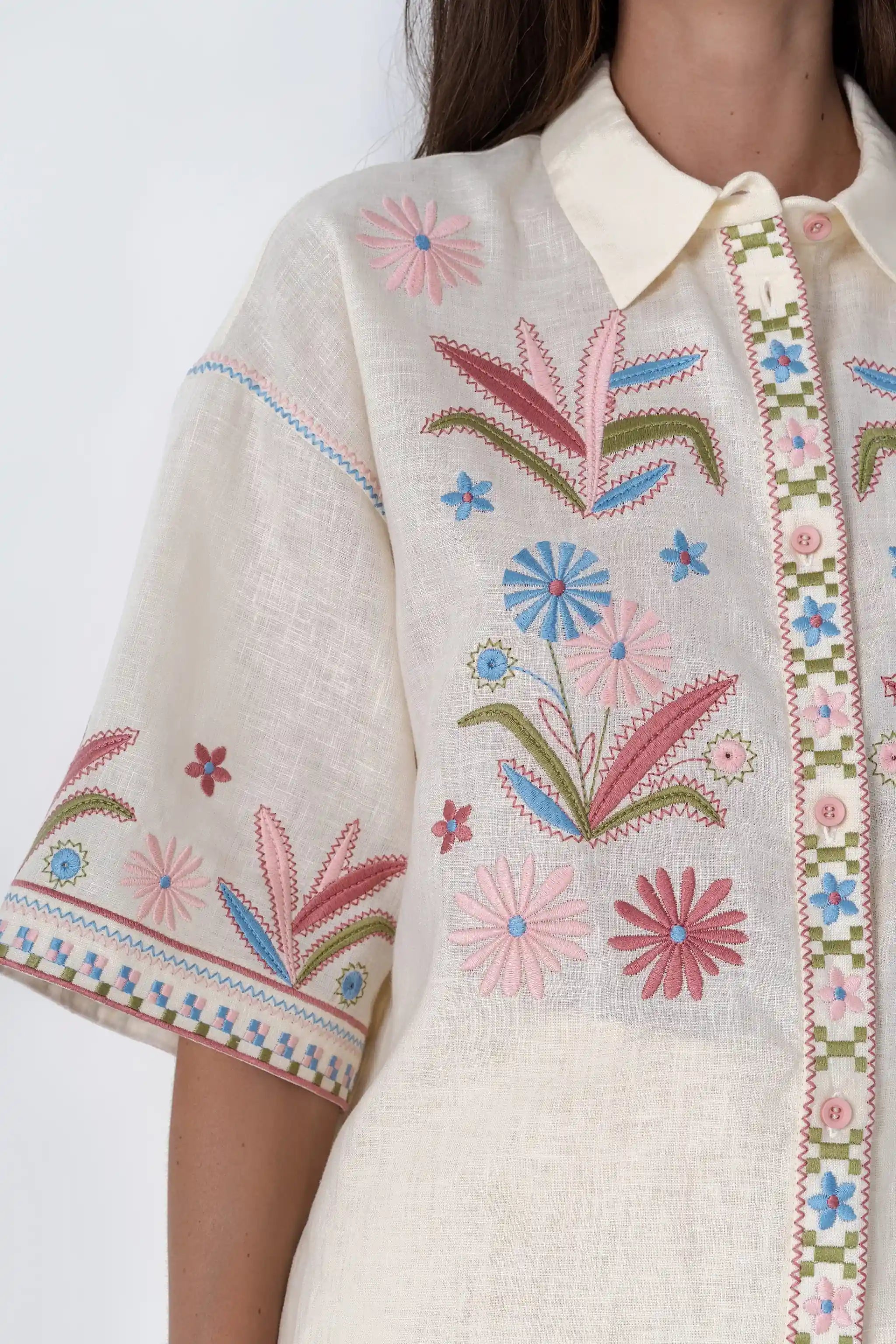 A woman modeling a cream-colored short-sleeve button-up shirt with colorful floral embroidery on the front and sleeves, paired with matching cream shorts and brown slide sandals, against a light gray studio background.