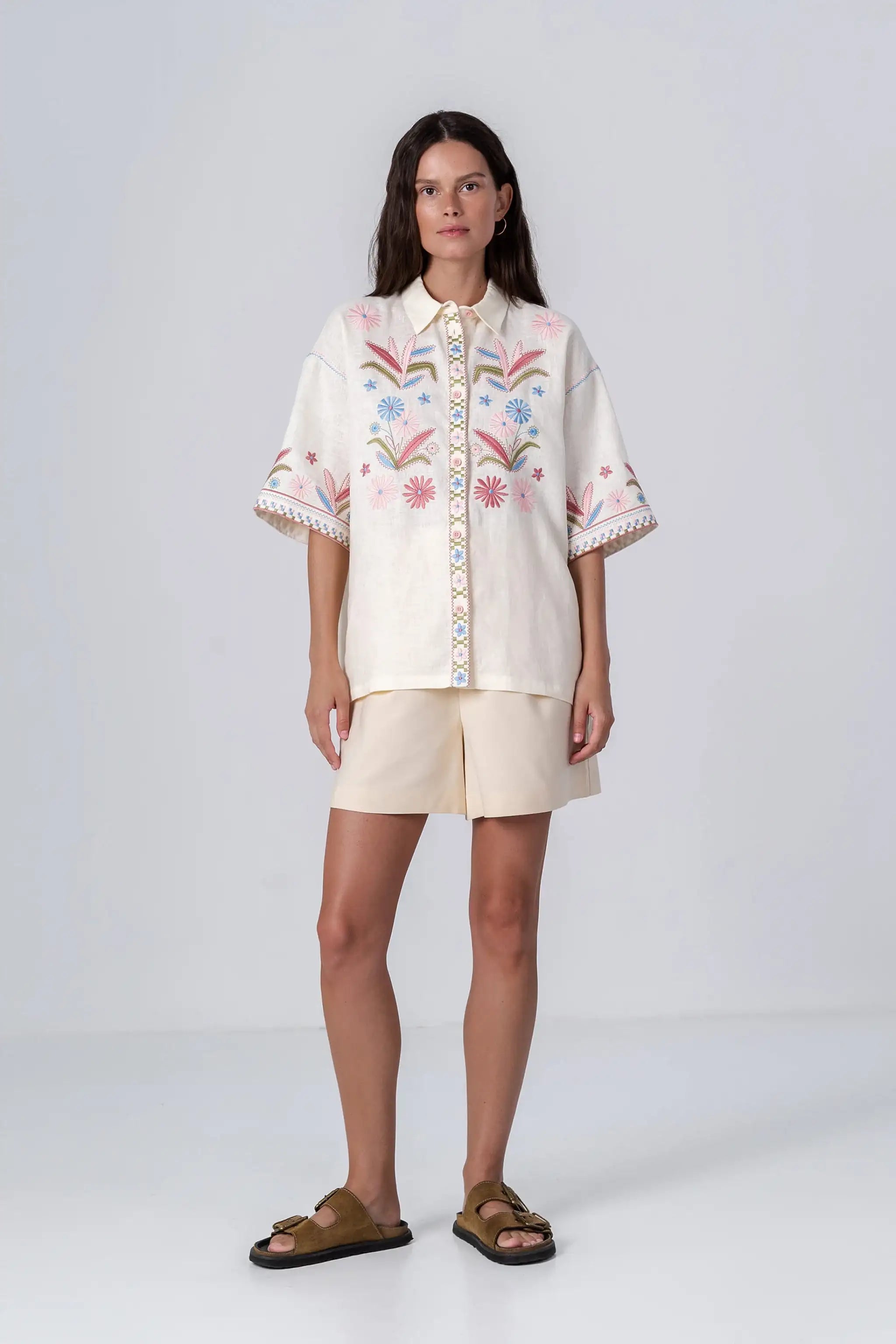 A woman modeling a cream-colored short-sleeve button-up shirt with colorful floral embroidery on the front and sleeves, paired with matching cream shorts and brown slide sandals, against a light gray studio background.