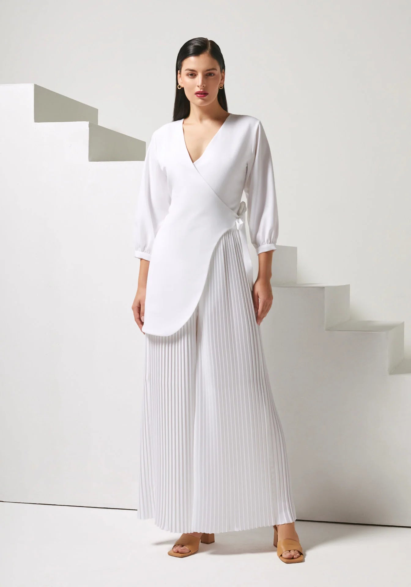 A woman modeling a white wrap-style long-sleeve top paired with matching white pleated wide-leg pants, standing in a minimalist white studio setting with geometric steps.