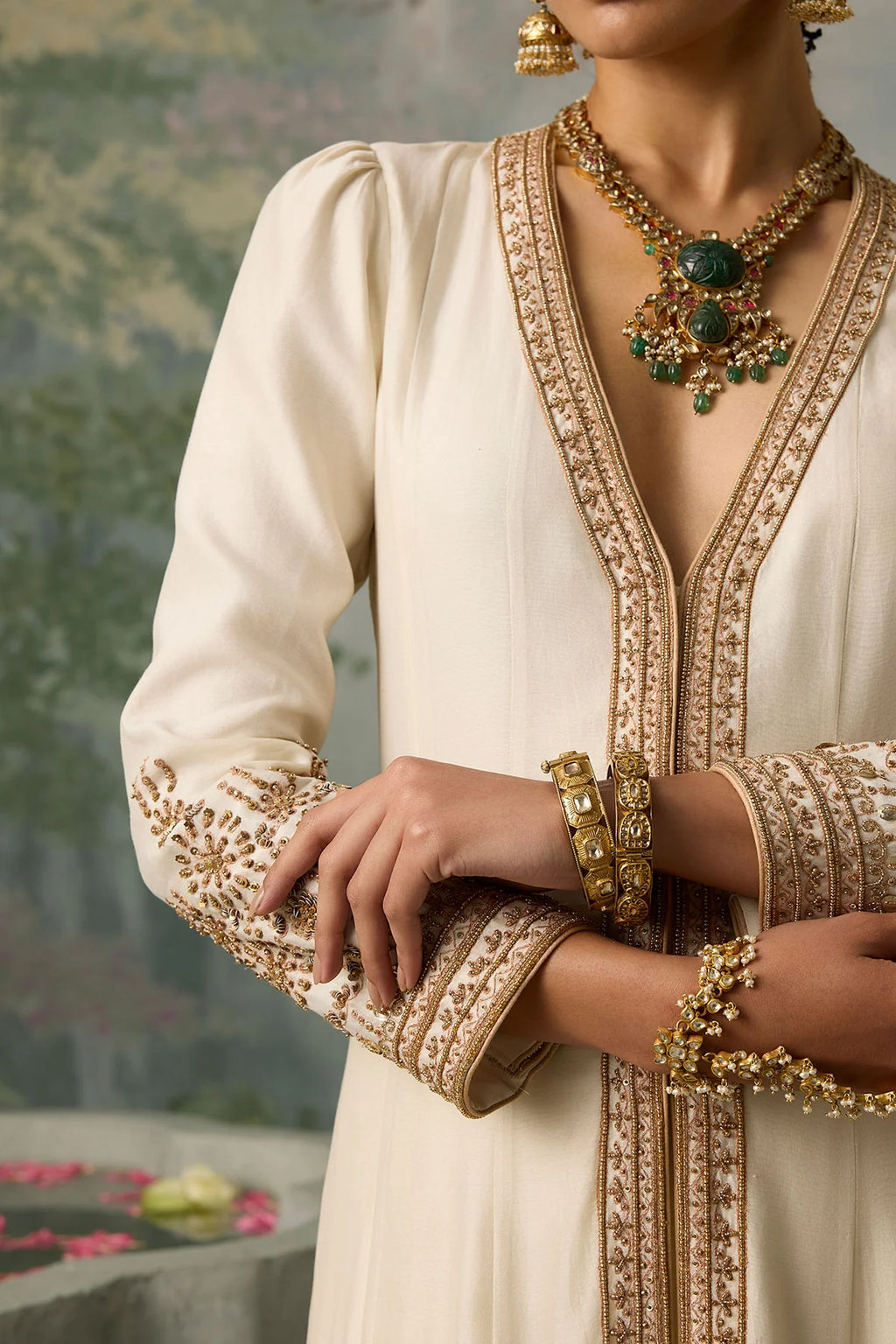Close-up of a person wearing an ornate beige outfit with gold embroidery, jewelry, and a blurred natural background.