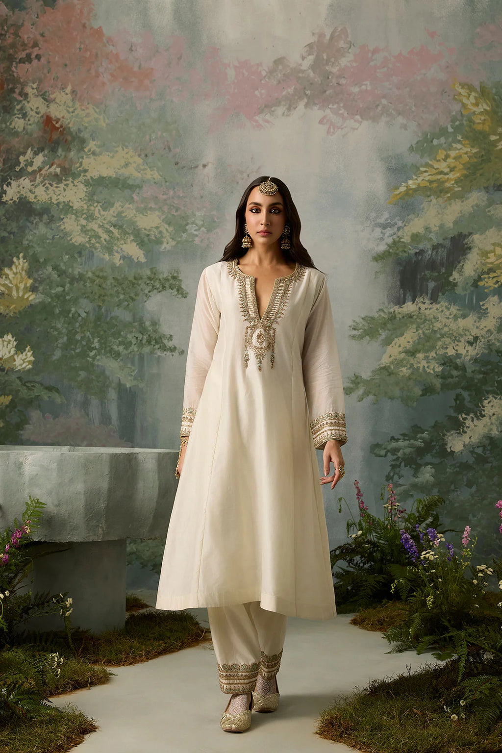 Woman in a white traditional outfit standing in a garden with a floral backdrop