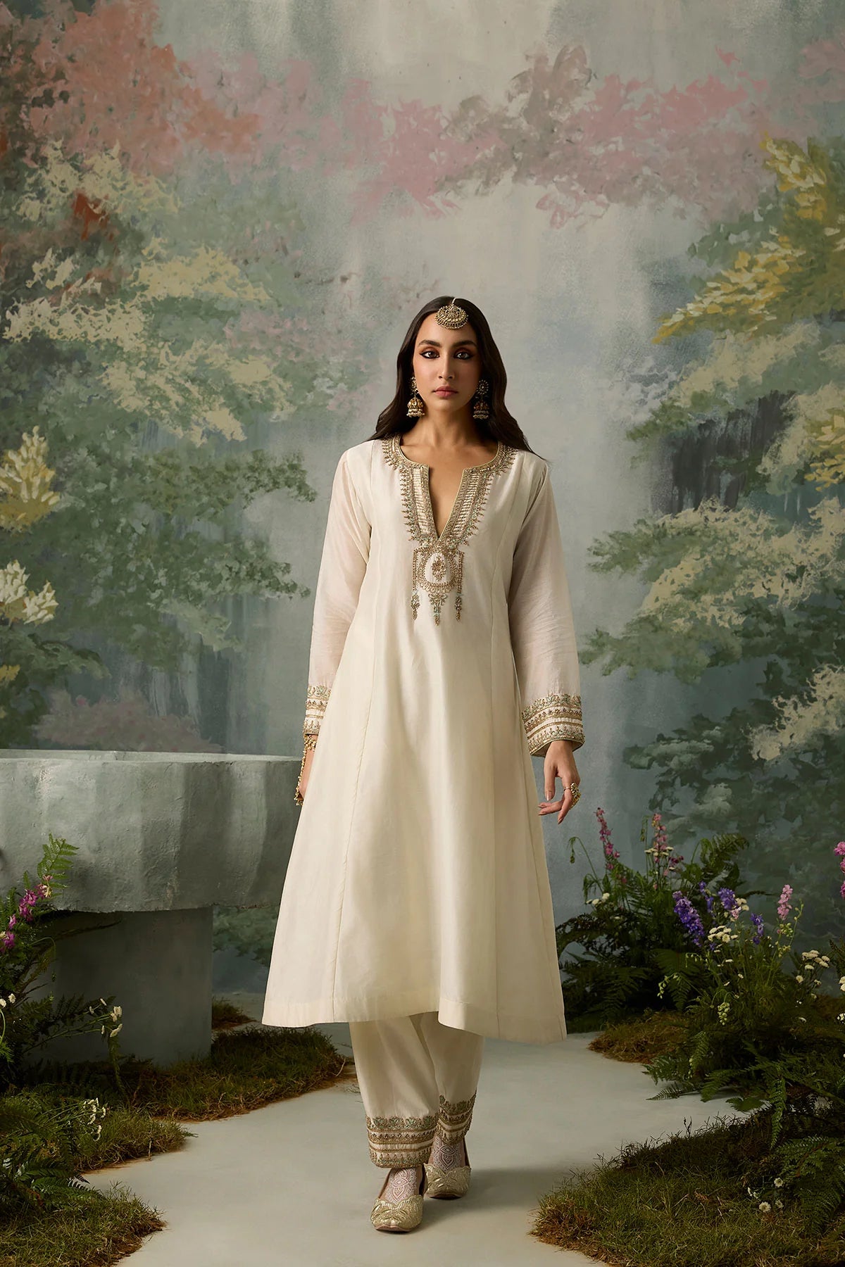 Woman in a white traditional outfit standing in a garden with a floral backdrop